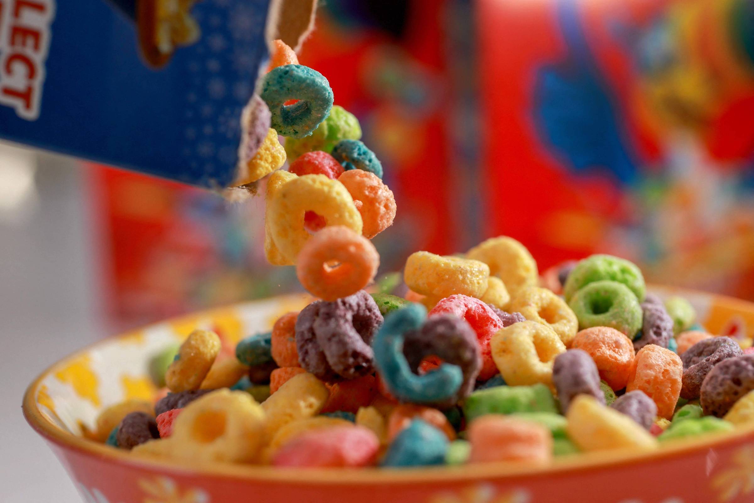 Uma caixa de cereal colorido está sendo despejada em uma tigela. O cereal é composto por várias formas e cores, incluindo anéis e pedaços em tons de amarelo, verde, azul, vermelho e roxo. A tigela é decorada com um padrão floral e está cheia de cereal. Ao fundo, há uma embalagem do cereal visível, com cores vibrantes.