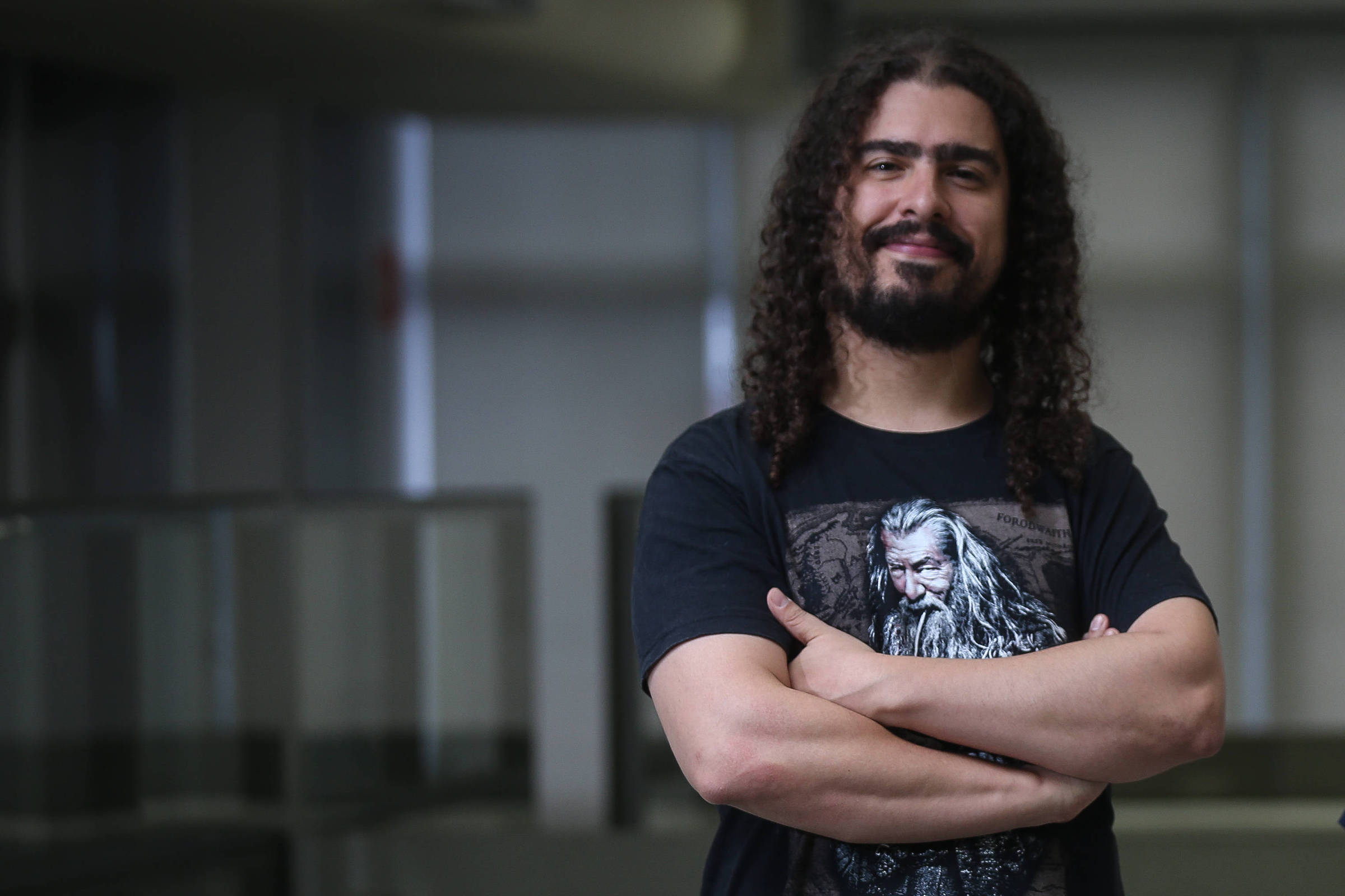 A imagem mostra dois homens em um ambiente de escritório. O homem à esquerda tem cabelo cacheado e usa uma camiseta preta com uma estampa. Ele está de braços cruzados e tem uma expressão confiante. O homem à direita está sentado em uma mesa, usando óculos e uma camiseta azul com uma estampa floral. O fundo é um espaço de escritório com mesas e janelas.