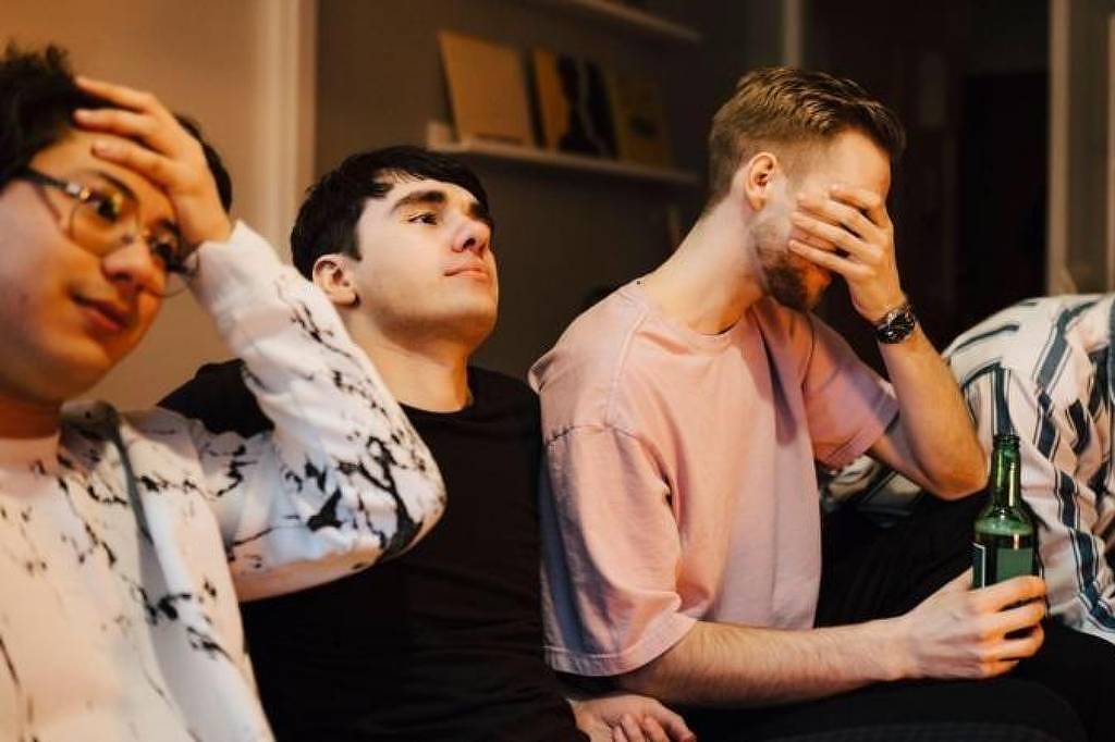 Cinco jovens homens estão sentados em um sofá, assistindo a algo. Dois deles estão com as mãos na cabeça, demonstrando preocupação ou frustração. Um terceiro homem está cobrindo o rosto com a mão, enquanto o quarto está segurando uma garrafa de bebida e sorrindo. O ambiente é interno, com uma decoração simples e moderna ao fundo.