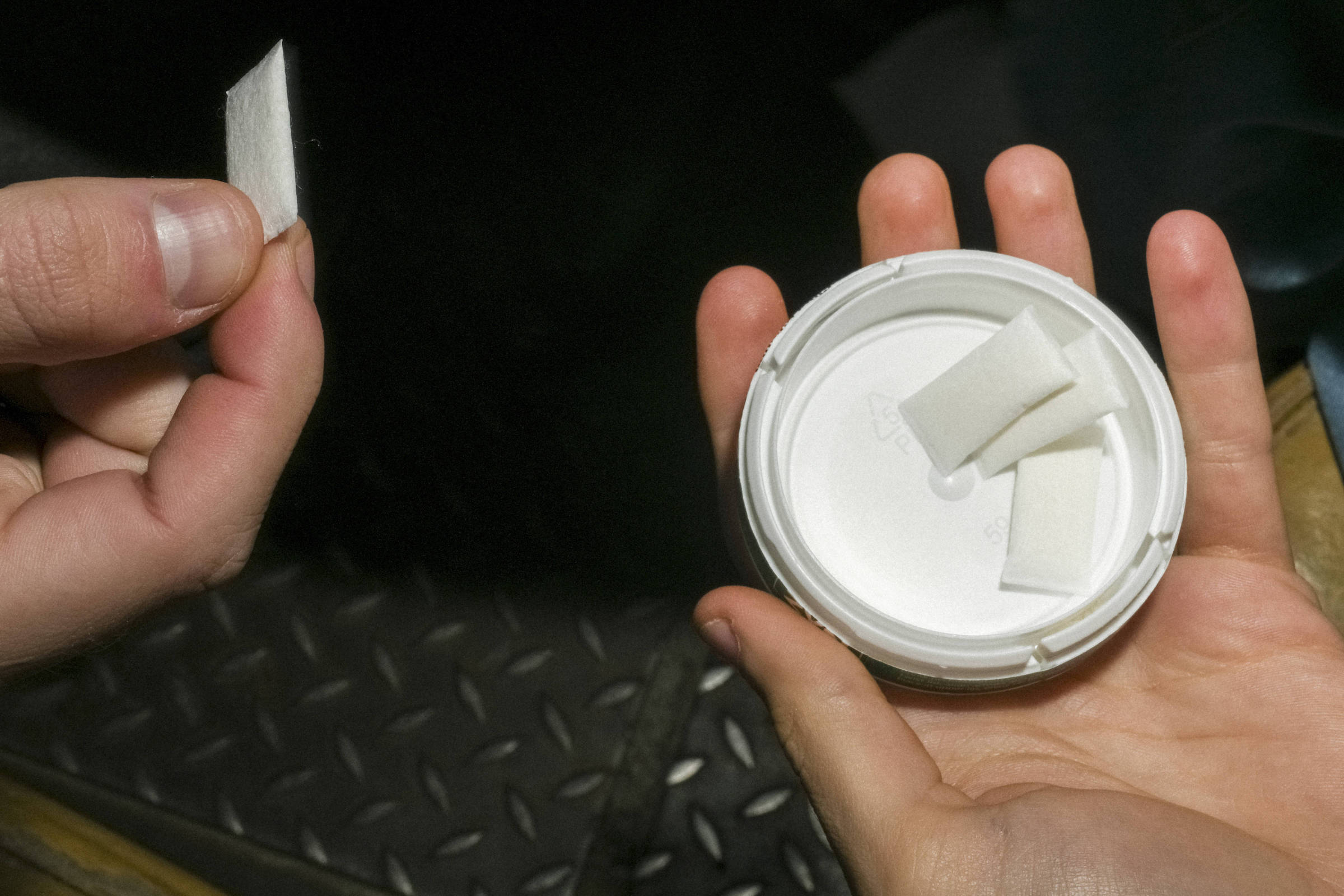 Uma mão segura um pequeno pedaço de papel enquanto a outra mão segura um recipiente branco com uma tampa. Dentro do recipiente, há vários pedaços de papel semelhantes ao que está sendo segurado. O fundo da imagem é escuro e apresenta uma superfície com um padrão de diamante.