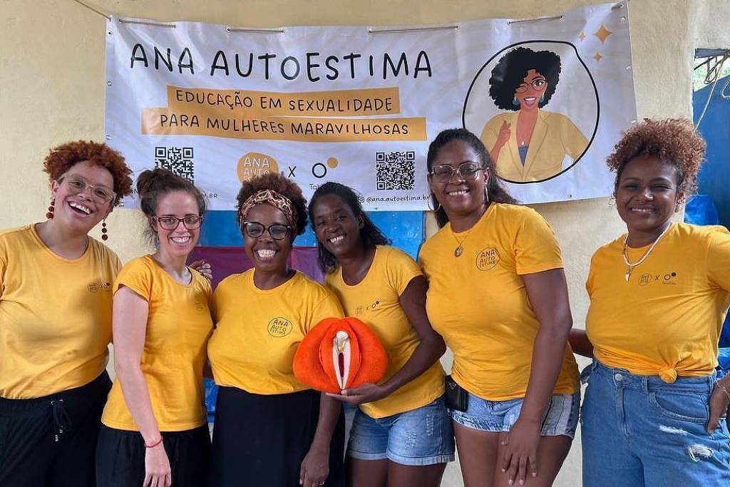 Seis mulheres estão posando para a foto em um evento. Todas estão vestindo camisetas amarelas. Elas estão sorrindo e algumas estão segurando um objeto laranja. Ao fundo, há um banner com o texto