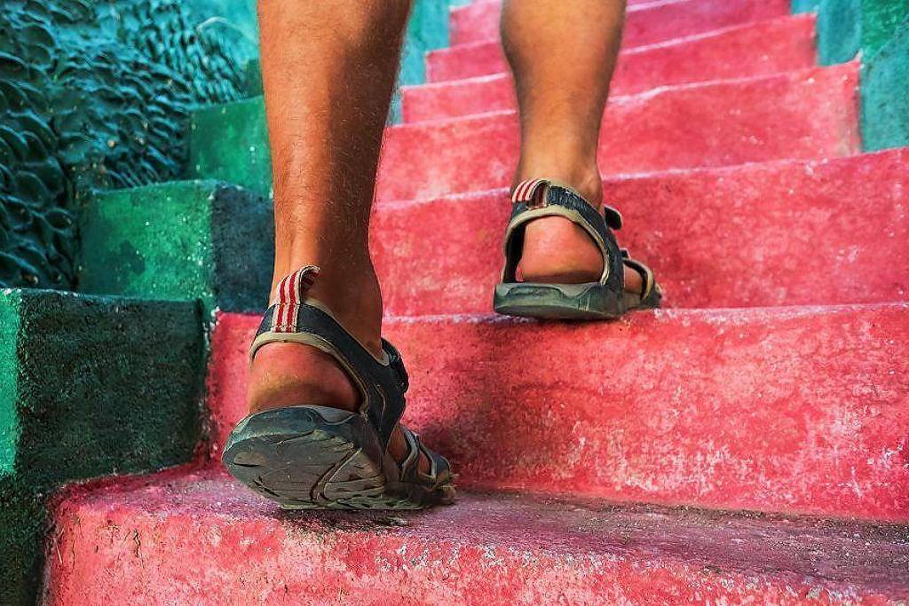 A imagem mostra uma pessoa subindo escadas coloridas. As escadas são pintadas em tons de rosa e verde, e a pessoa está usando sandálias. A parte inferior das pernas é visível, destacando a textura das escadas e o calçado da pessoa.