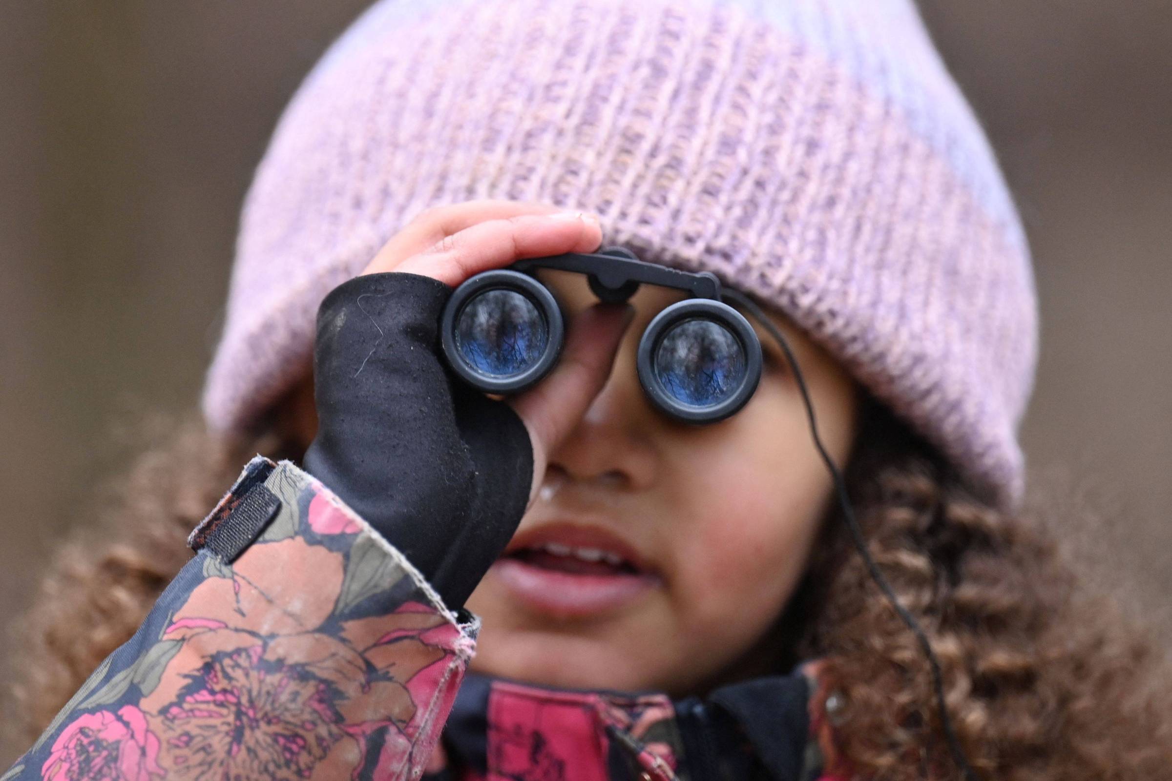 Uma criança com cabelo cacheado e pele clara está usando binóculos. Ela está vestindo um casaco colorido com estampas florais e um gorro de lã rosa. A criança parece estar concentrada enquanto observa algo à sua frente.