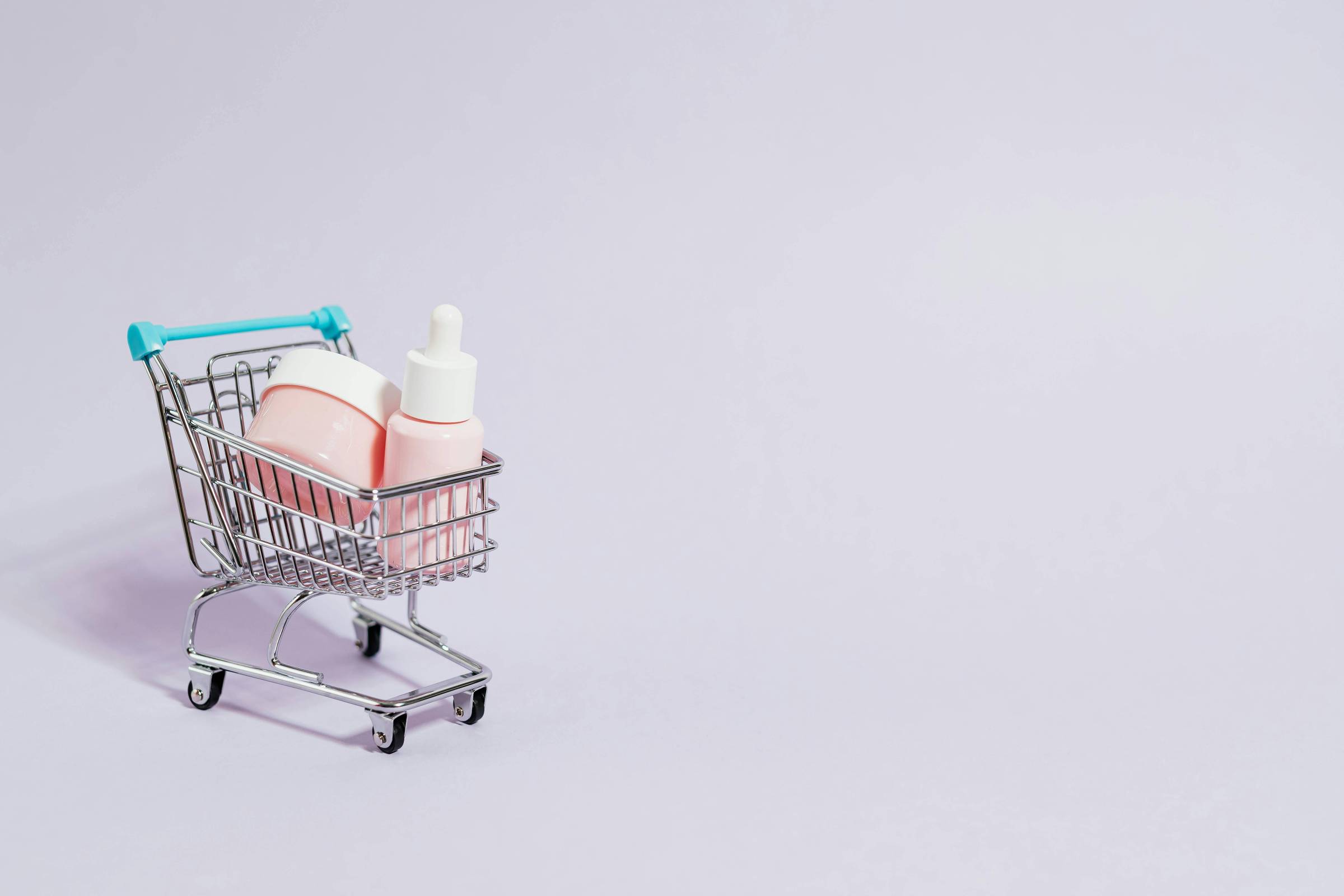 Um pequeno carrinho de compras de metal com dois frascos de produtos de beleza. Um dos frascos é um pote rosa com tampa branca e o outro é um frasco rosa com conta-gotas branco. O fundo é de uma cor lilás clara.