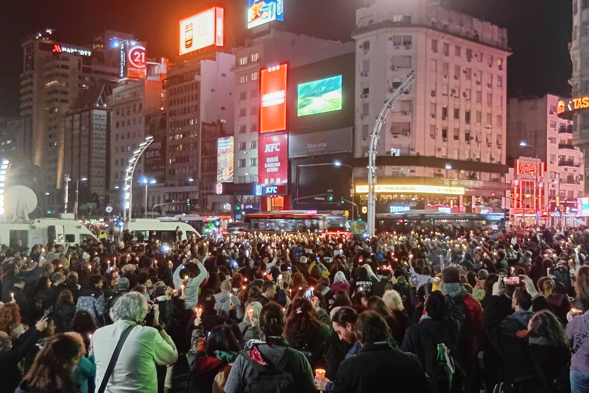 Um grupo de pessoas segura velas e lanternas no meio de uma avenida. Há uma grande quantidade de luzes brilhantes e anúncios em faixas nas fachadas dos edifícios. A iluminação é artificial, composta principalmente por luzes de anúncios e vitrines dos edifícios. A atmosfera é vibrante, dinâmica e moderna, capturando a energia e o movimento constantes da área.