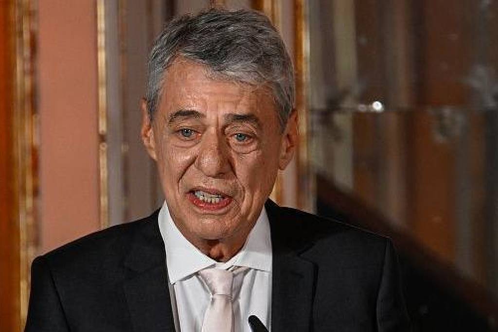 Um homem de cabelo grisalho e curto, vestindo um terno escuro e uma gravata clara, está falando ao microfone. Ele parece estar em um evento formal, com um fundo que sugere um ambiente elegante. A expressão facial do homem é séria e concentrada.