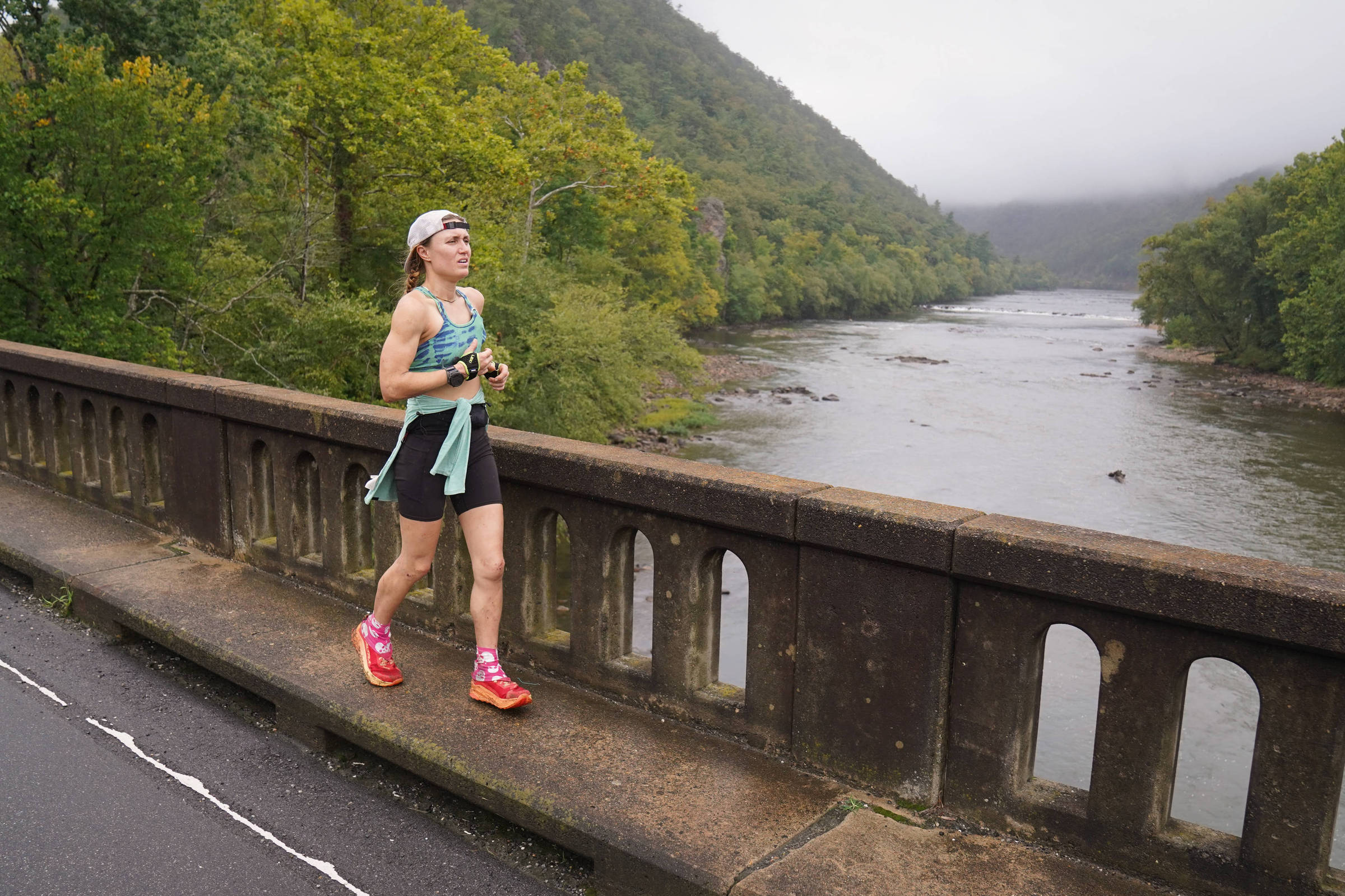 Uma mulher está correndo ao longo de uma ponte de pedra, com um rio calmo à sua esquerda. O cenário é cercado por árvores verdes e uma montanha ao fundo, coberta por névoa. A corredora usa uma blusa azul clara e shorts pretos, além de tênis rosa. O clima parece nublado e úmido.