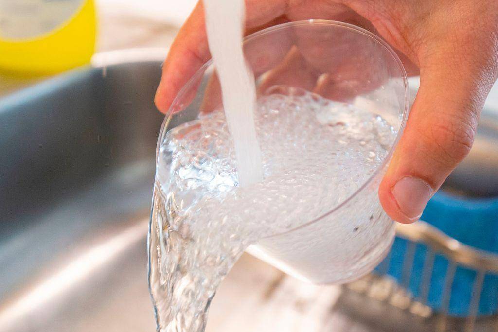 A imagem mostra uma mão segurando um copo transparente enquanto água está sendo despejada nele. O copo está quase cheio, e a água é clara e borbulhante. Ao fundo, pode-se ver uma pia de cozinha com um escorredor azul e um objeto amarelo não identificado.