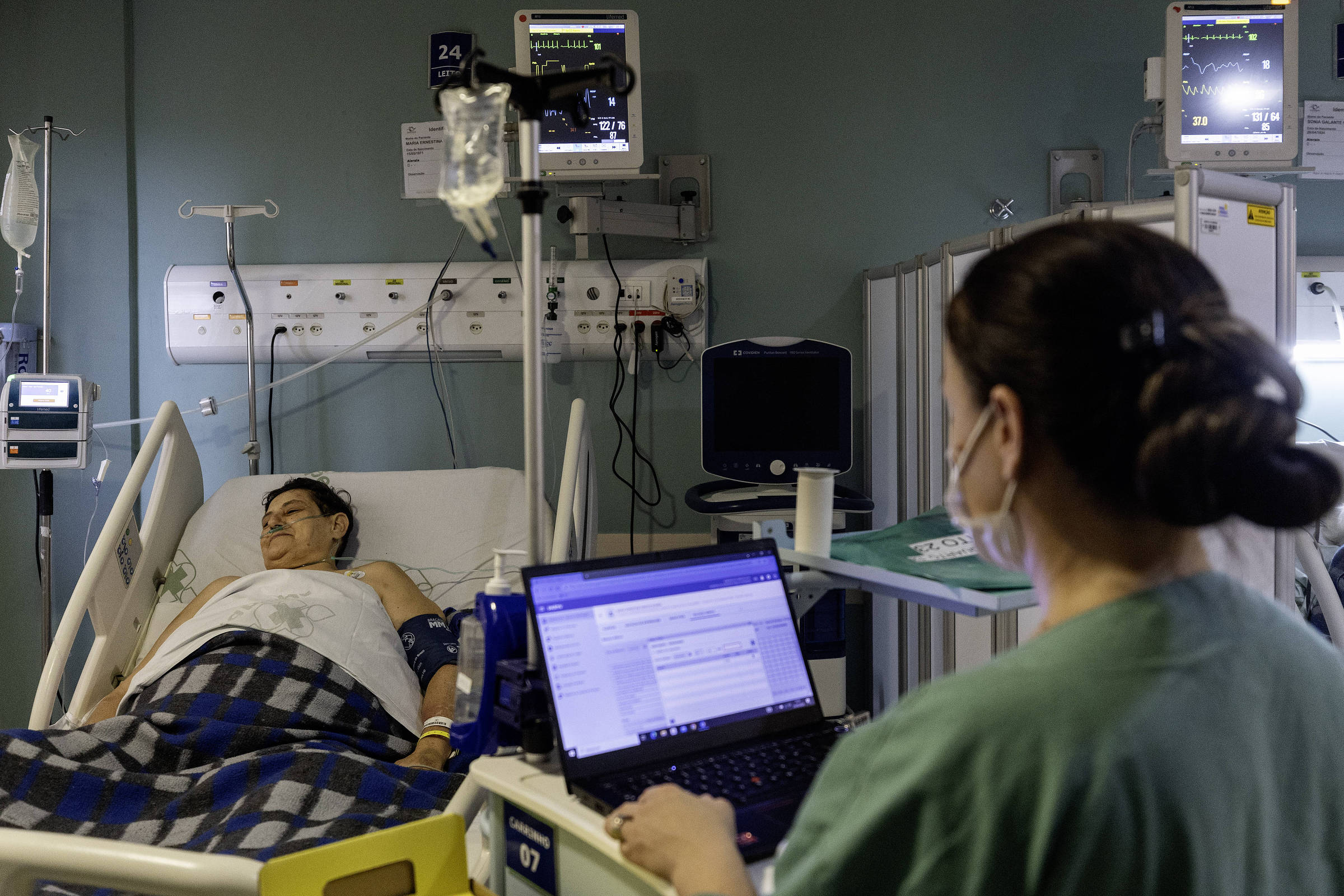 A imagem mostra um paciente deitado em uma cama de hospital, coberto com um cobertor xadrez. Ao lado do leito, uma profissional de saúde, usando máscara, está sentada em uma cadeira, trabalhando em um laptop. No fundo, há monitores médicos e equipamentos hospitalares visíveis.
