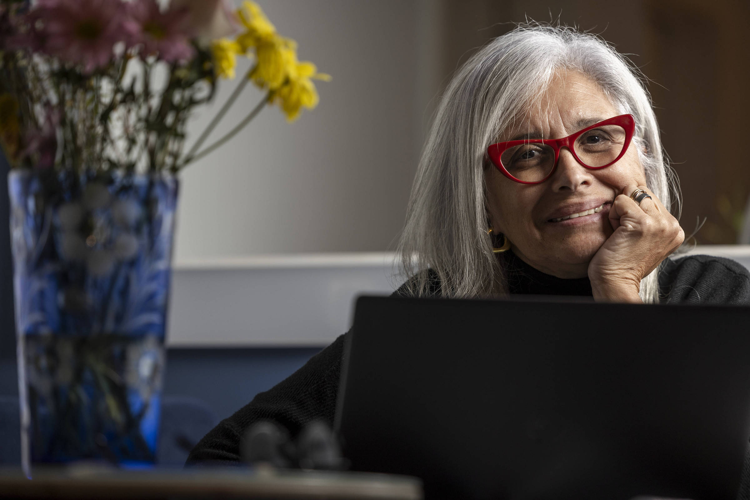 Uma mulher de cabelos longos e grisalhos, usando óculos vermelhos, está sentada em frente a um laptop. Ela sorri levemente enquanto apoia o queixo na mão, parecendo pensativa. Ao fundo, há um vaso de flores coloridas, incluindo flores amarelas. O ambiente é iluminado e acolhedor.