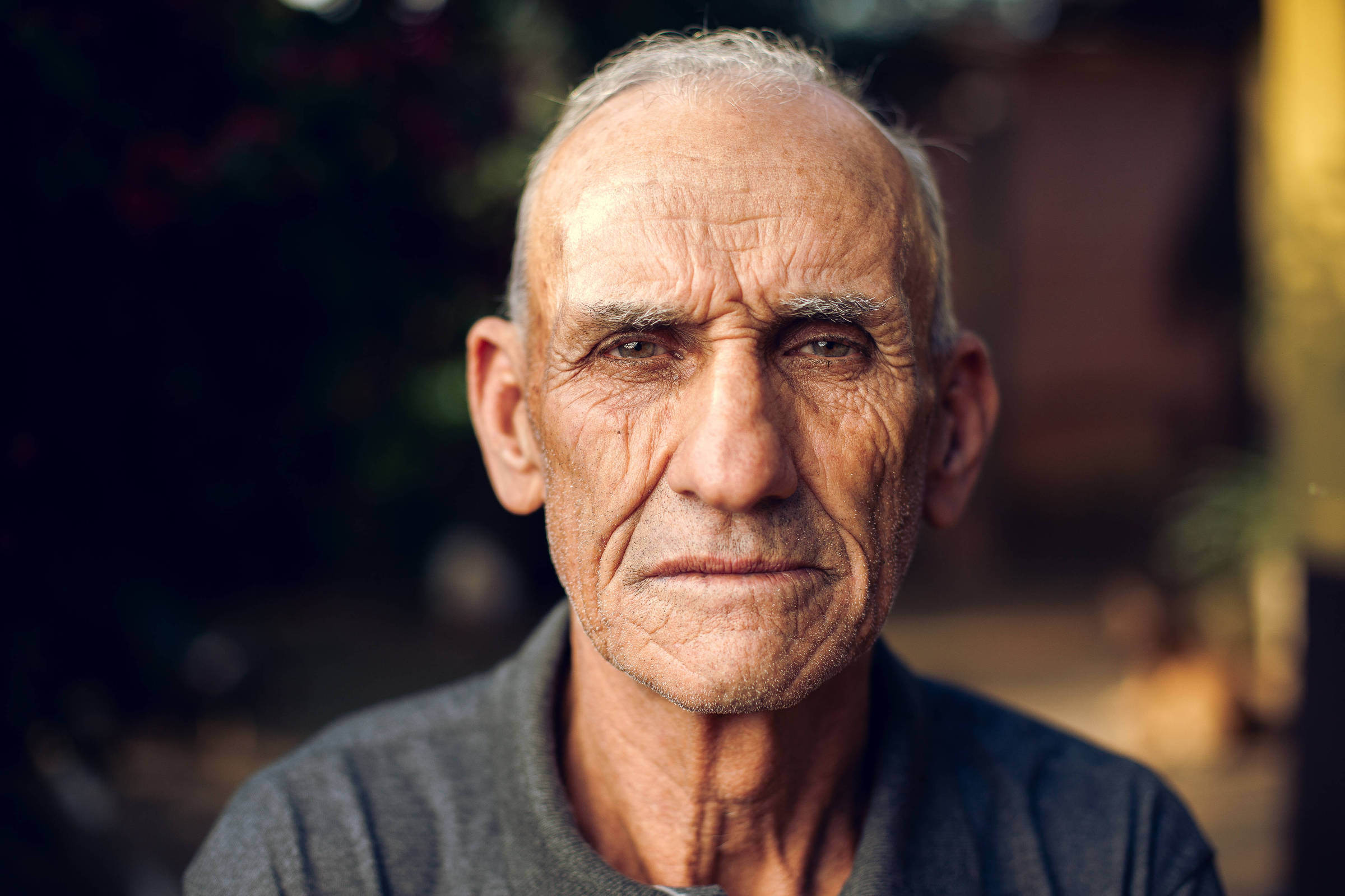 Um homem idoso está olhando diretamente para a câmera. Ele tem cabelo grisalho e uma expressão séria. O fundo é desfocado, com algumas plantas visíveis, sugerindo um ambiente ao ar livre. O homem está vestindo uma camisa de gola cinza.