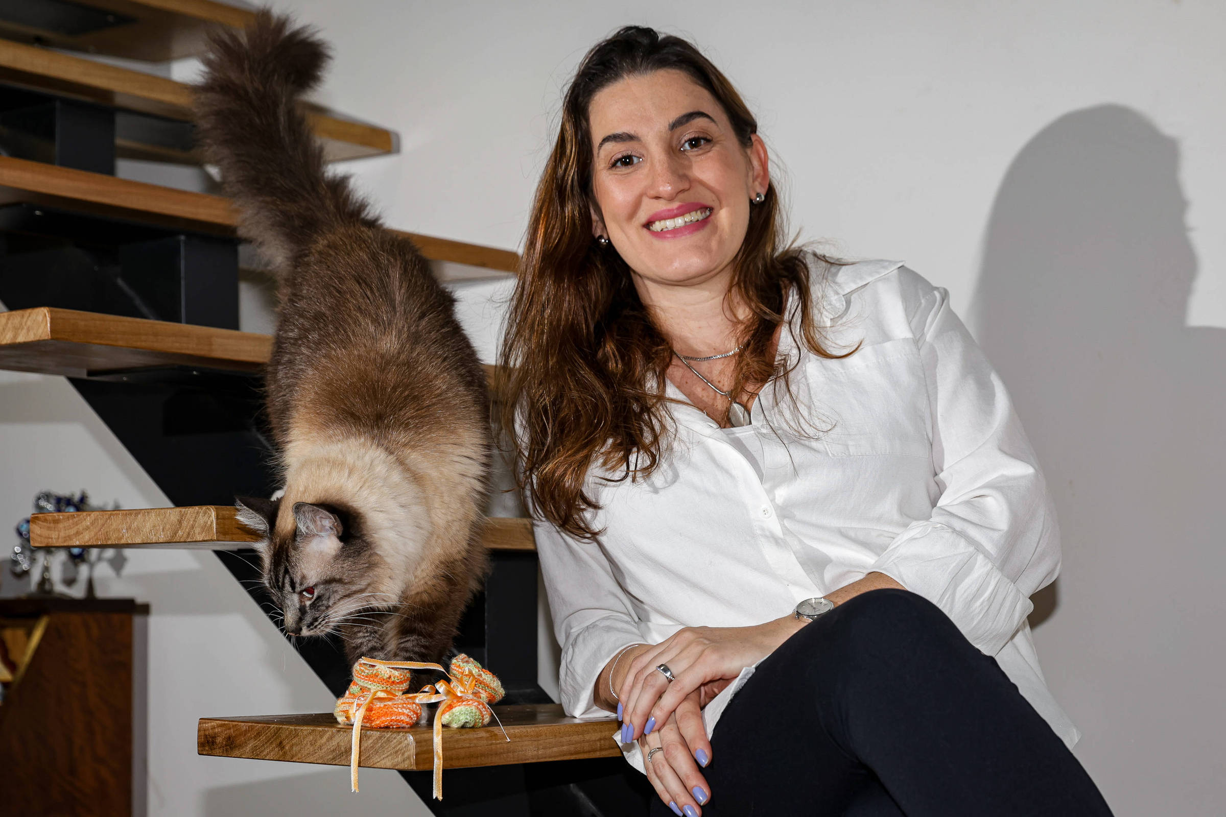 A imagem mostra uma mulher sorridente sentada em uma escada de madeira. Ela está vestindo uma camisa branca e calças escuras. Ao seu lado, um gato de pelagem clara está caminhando em direção a um objeto decorativo que parece ser um par de sapatos de brinquedo. O fundo é uma parede clara e a escada tem degraus expostos.
