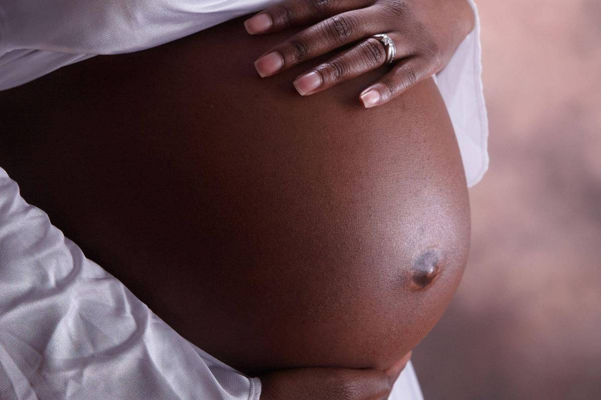 A imagem mostra uma barriga de grávida, com a pele de tom escuro. As mãos de uma pessoa estão posicionadas sobre a barriga, uma delas usando um anel. O fundo é desfocado, com tons suaves e neutros.
