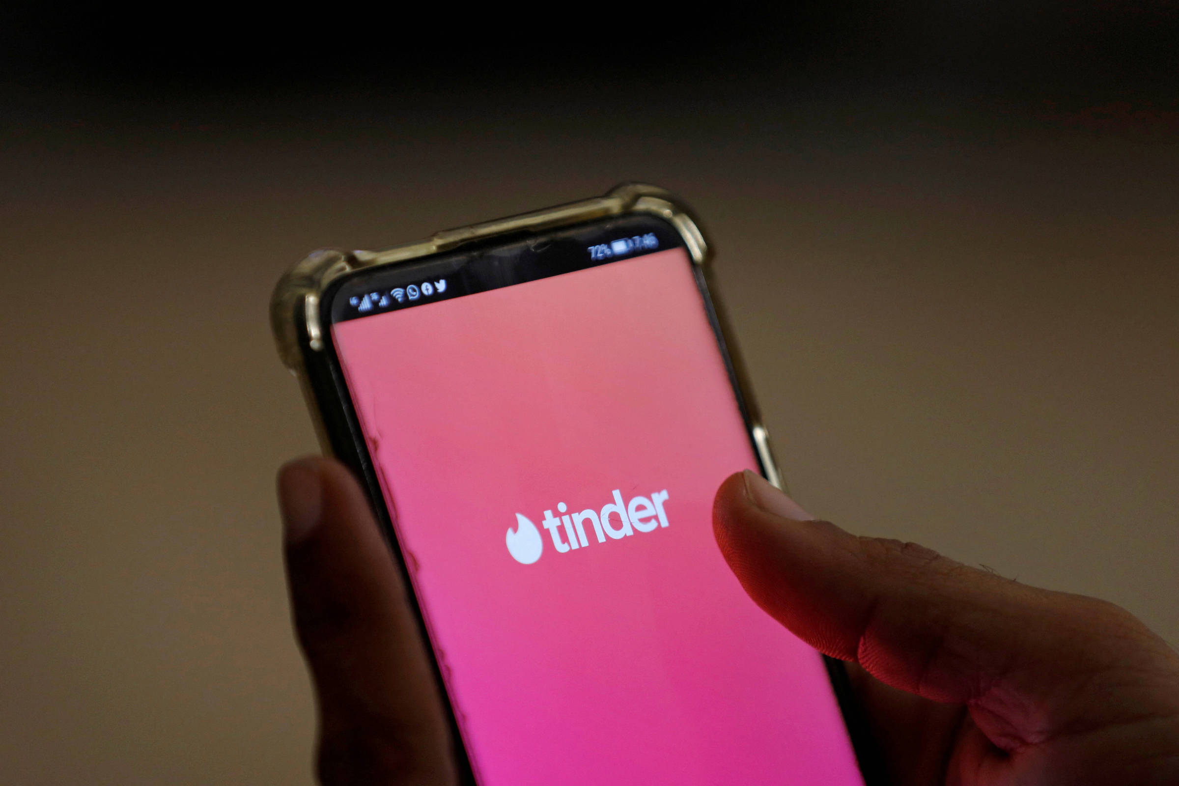 A imagem mostra um smartphone segurado por uma mão, exibindo a tela inicial do aplicativo Tinder. O fundo da tela é de um gradiente rosa, com o logotipo do Tinder em branco no centro. A parte superior da tela mostra ícones de sinal e bateria.