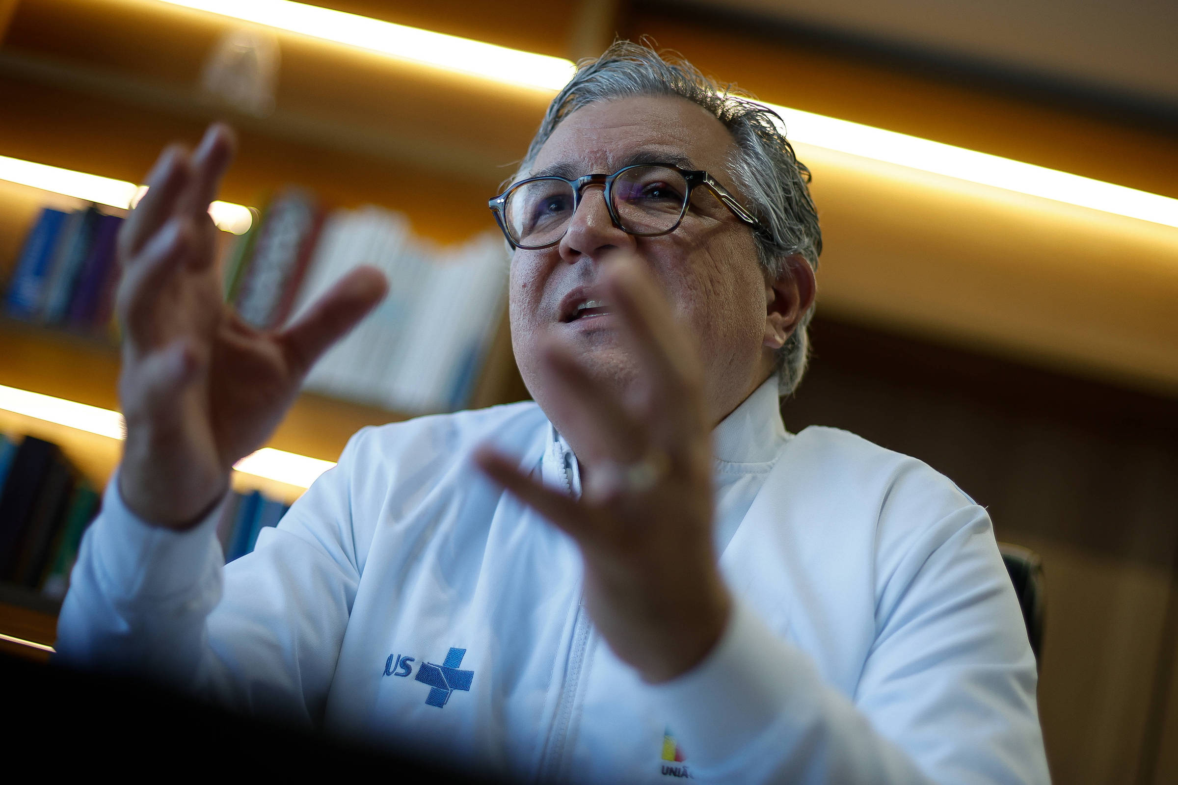 Um homem com cabelo grisalho e óculos está gesticulando enquanto fala. Ele usa uma camisa branca com um logotipo visível. Ao fundo, há prateleiras com livros. A iluminação é suave, destacando o rosto do homem e suas expressões.
