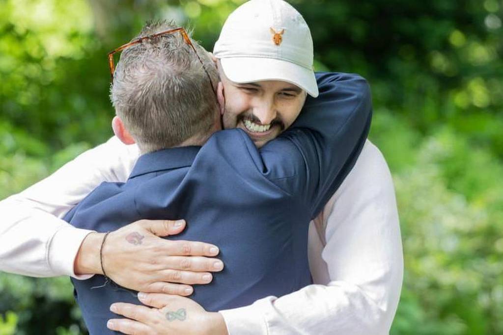 Na imagem, duas pessoas estão se abraçando em um ambiente ao ar livre, cercado por vegetação. Uma das pessoas está vestindo uma camisa escura e a outra está usando uma camiseta clara e um boné. Ambas parecem felizes e sorrindo durante o abraço.