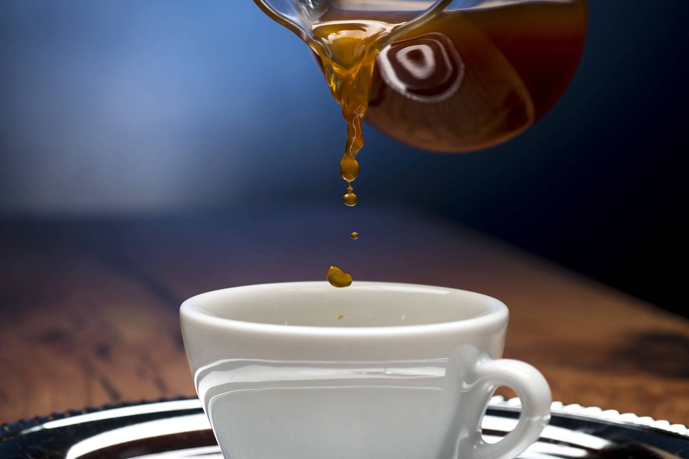 Uma mão está segurando um recipiente de vidro, despejando um líquido marrom em uma xícara branca. A xícara está posicionada sobre uma bandeja prateada, e o fundo é desfocado, com uma iluminação suave que destaca a cena.