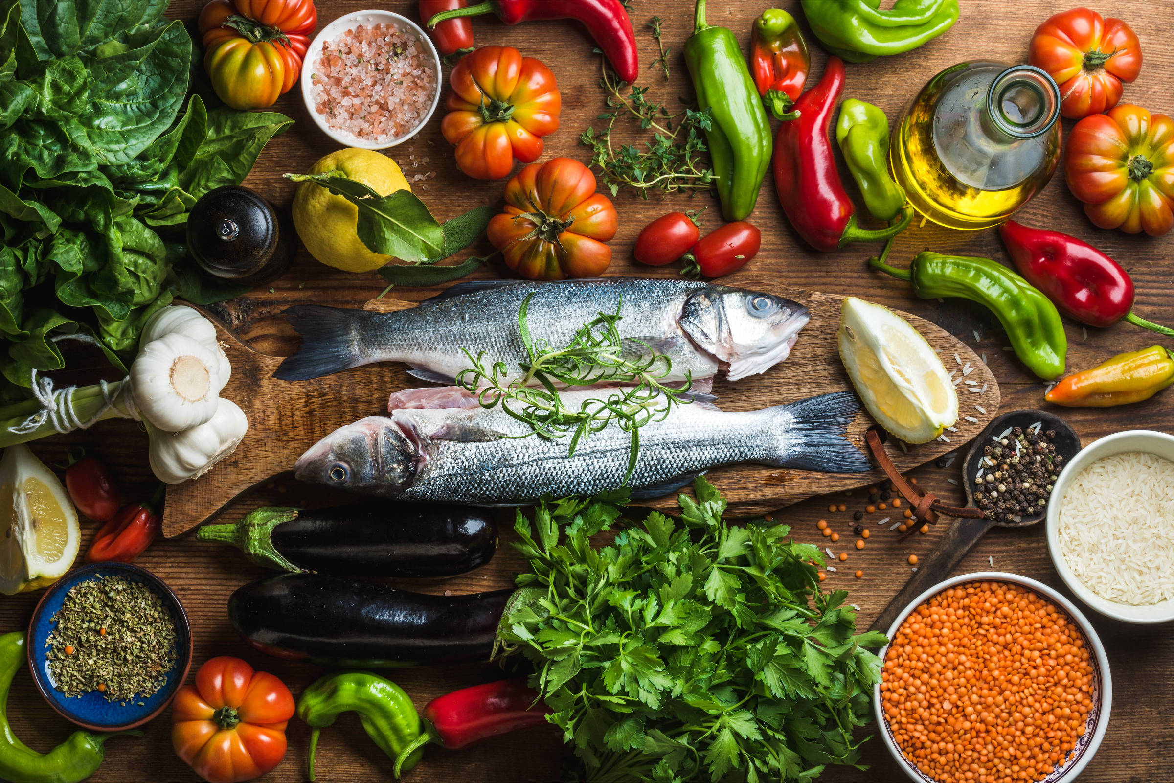 A imagem apresenta uma variedade de ingredientes frescos dispostos sobre uma superfície de madeira. No centro, há dois peixes inteiros, cercados por ervas, como salsinha e espinafre. Também estão presentes tomates de diferentes cores, pimentões verdes e vermelhos, limões, alho, azeite, sal, lentilhas, arroz e berinjela. A composição é colorida e vibrante, refletindo uma seleção saudável de alimentos.