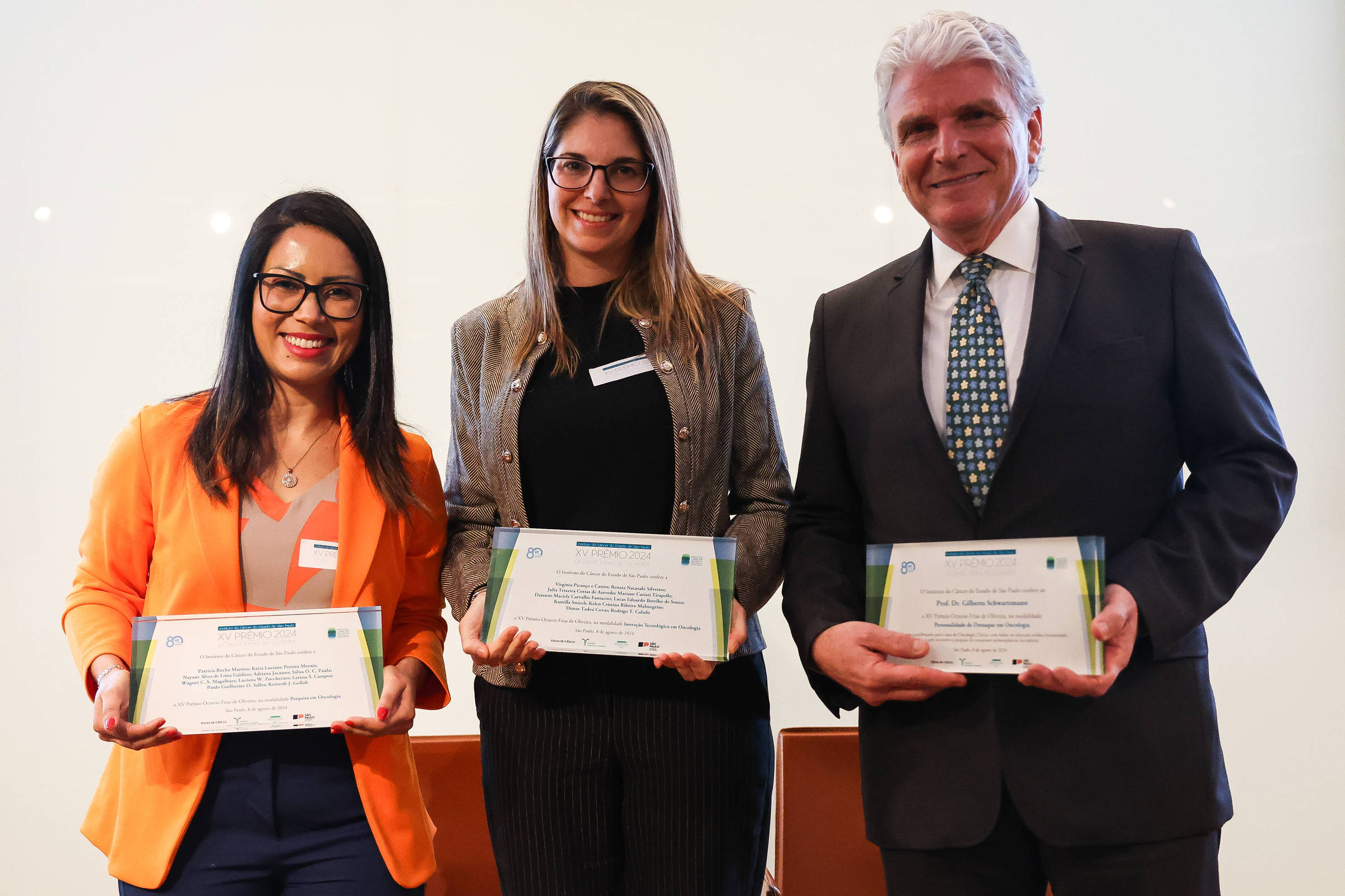 Três pessoas estão em pé, segurando certificados em uma cerimônia de premiação. A primeira pessoa à esquerda é uma mulher com óculos e cabelo longo, vestindo uma blusa preta e um blazer laranja. A segunda pessoa, no centro, é uma mulher com cabelo liso e óculos, vestindo uma blusa preta e calças listradas. A terceira pessoa à direita é um homem com cabelo grisalho, usando um terno escuro e uma gravata com padrões. Todos estão sorrindo e parecem felizes com a premiação.
