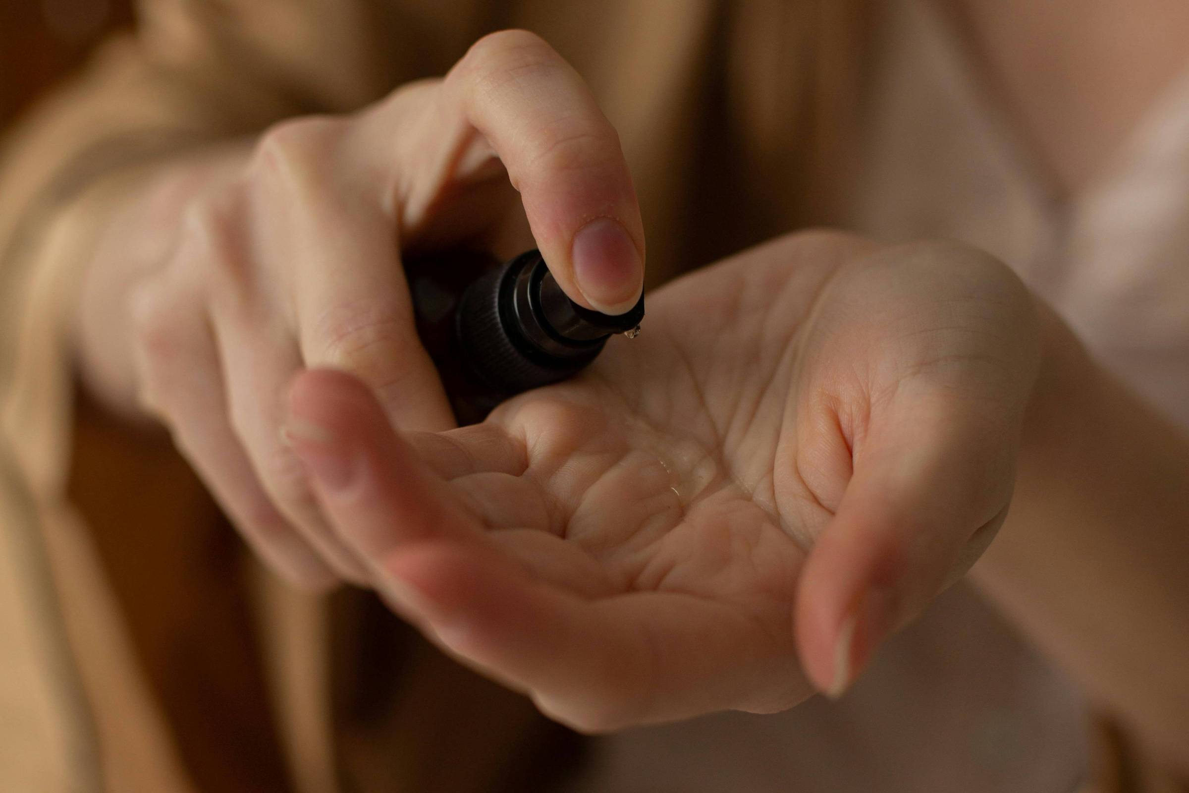 A imagem mostra uma pessoa aplicando um óleo em uma das mãos. A mão esquerda segura um frasco de óleo, enquanto a mão direita está aberta, recebendo o produto. A pessoa está vestindo uma roupa clara e um robe, e o fundo é desfocado, sugerindo um ambiente aconchegante.