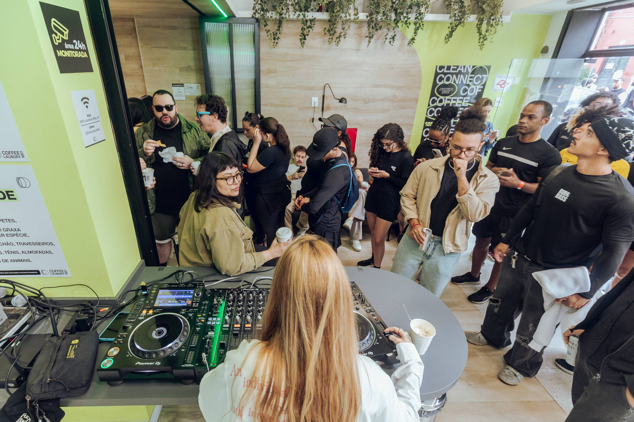 A imagem mostra um evento em um espaço moderno, onde uma DJ está tocando música em uma mesa de som. O público, composto por várias pessoas, está em pé, algumas segurando copos, enquanto outras interagem entre si. O ambiente é iluminado e decorado com plantas, e há uma lousa com texto visível ao fundo. A DJ tem cabelo longo e liso, e está de costas para a câmera, concentrada em sua performance.