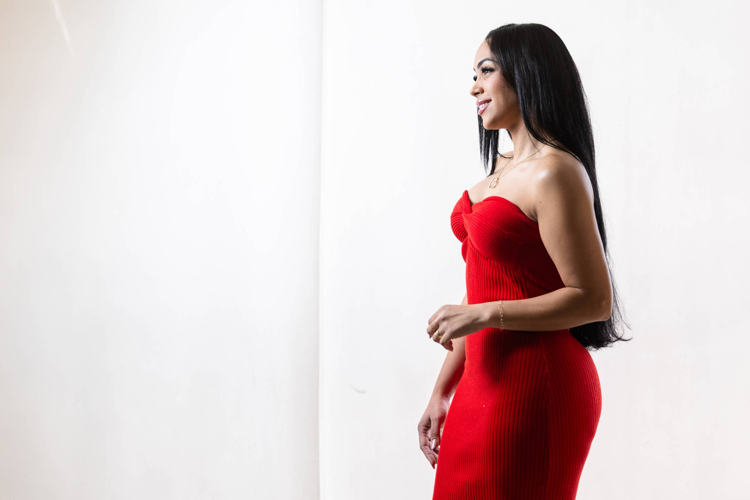 A imagem mostra uma mulher de perfil, usando um vestido vermelho justo e sem mangas. Ela tem cabelo longo e liso, e está em um ambiente com fundo claro. A mulher parece estar sorrindo levemente e está em uma pose elegante, com uma das mãos na cintura.