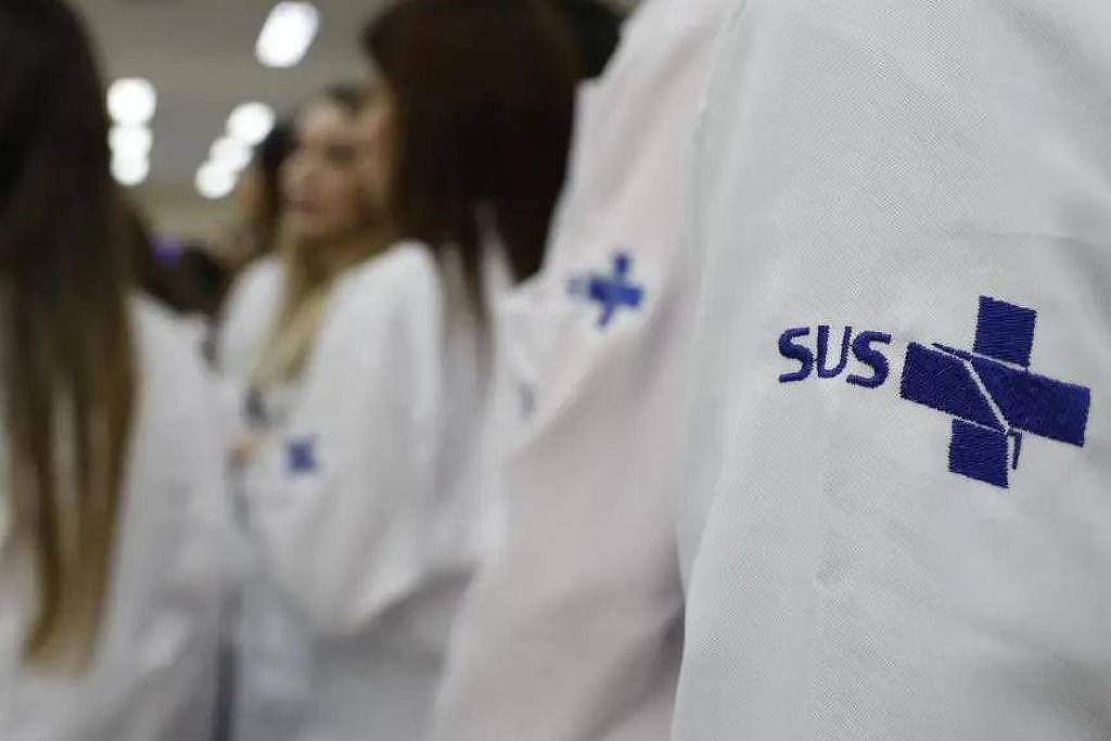 A imagem mostra um grupo de pessoas de costas, vestindo jalecos brancos. Em destaque, um jaleco à frente exibe o logo do SUS (Sistema Único de Saúde) em azul, que consiste em um texto