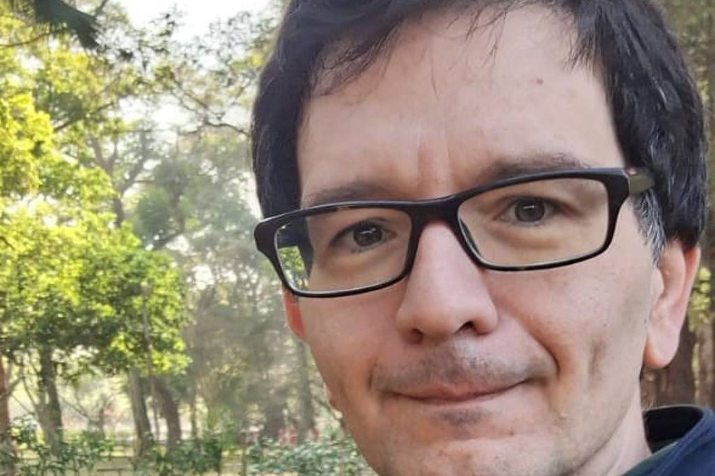 Um homem está posando para a foto em um ambiente natural, cercado por árvores e vegetação. Ele usa uma jaqueta escura e óculos. O fundo é desfocado, mas é possível ver folhas secas no chão e a luz do sol filtrando entre as árvores.