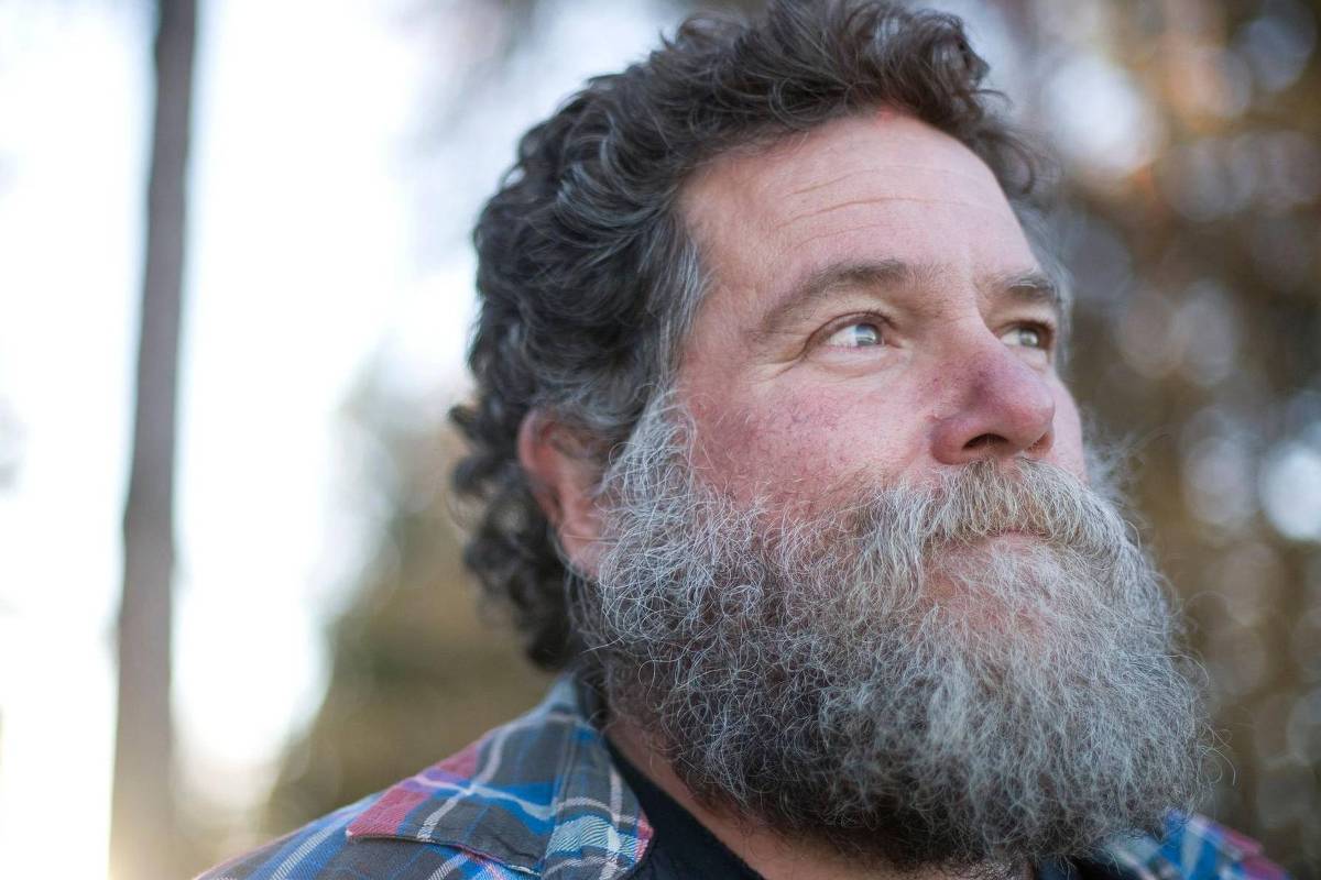 Um homem com barba espessa e cabelo encaracolado, olhando para o lado. Ele está usando uma camisa xadrez colorida e o fundo é desfocado, com árvores ao fundo. A luz é suave, sugerindo um ambiente natural.