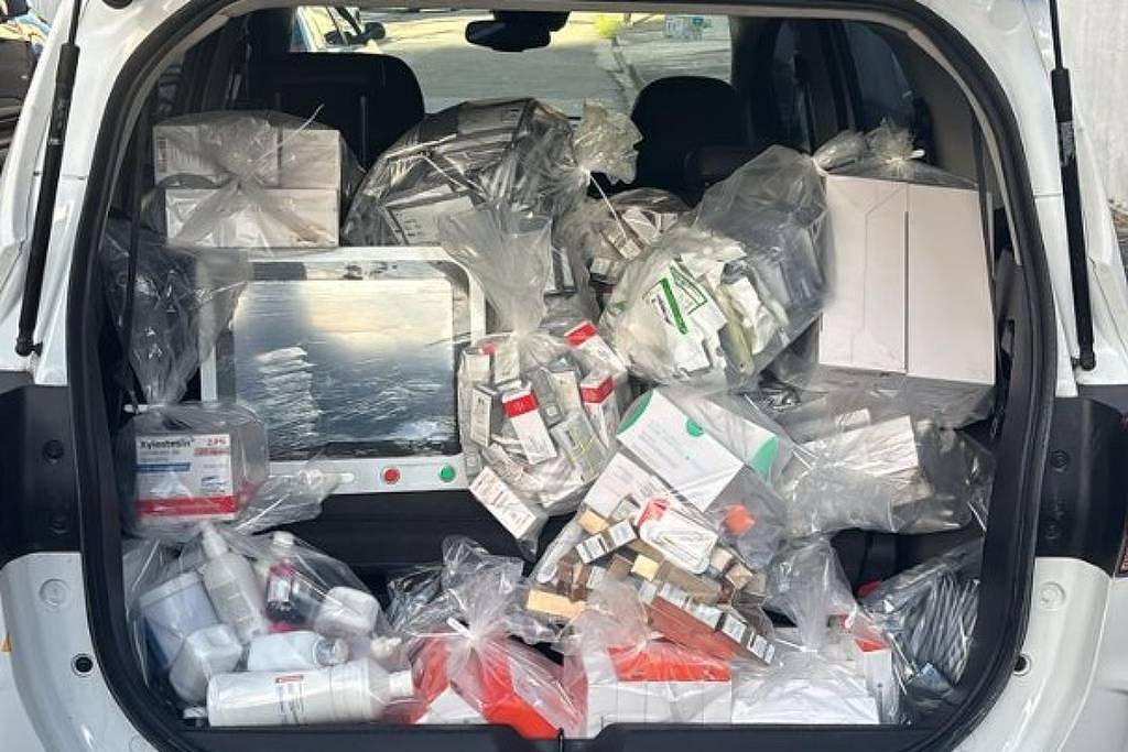 A imagem mostra o porta-malas de um carro branco cheio de sacolas plásticas e caixas. Os itens parecem ser produtos variados, possivelmente de supermercado ou farmácia, organizados de forma desordenada. Ao fundo, é possível ver prédios e uma área urbana.