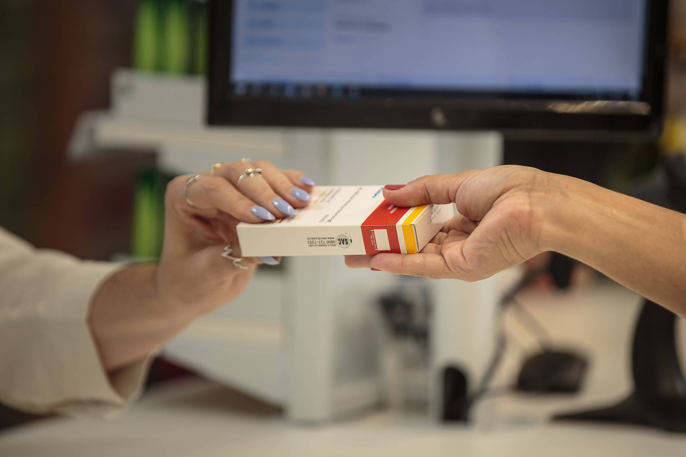 Uma mão está passando uma caixa de medicamento ou produto para outra mão em um balcão. A mão que recebe o produto tem unhas pintadas de azul e a mão que entrega é de uma pessoa com anéis. Ao fundo, há um computador com uma tela visível, mas sem informações legíveis.