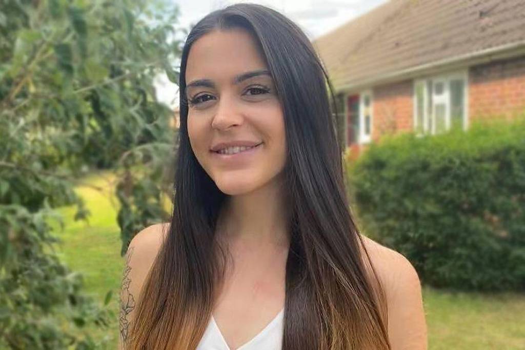 Uma jovem mulher está sorrindo em um ambiente ao ar livre. Ela tem cabelos longos e lisos, com as pontas mais claras. Ao fundo, há uma casa com telhado de duas águas e uma área verde com árvores e arbustos. A mulher está vestindo uma blusa branca e parece estar em um dia ensolarado.