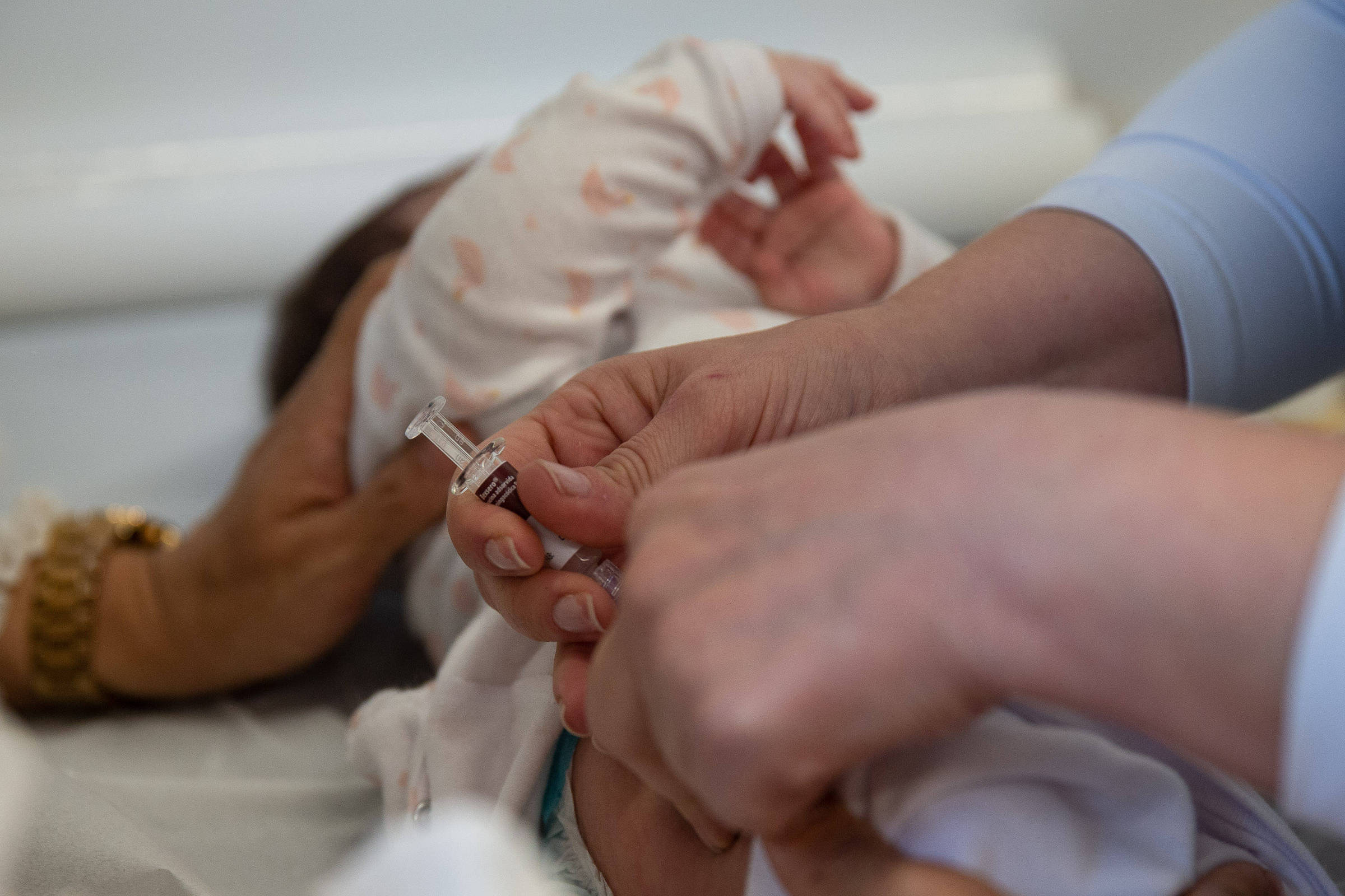 A imagem mostra um bebê deitado em uma superfície clara, enquanto uma pessoa está segurando uma seringa e se preparando para aplicar uma vacina. A mão da pessoa que aplica a vacina é visível, assim como a mão de outra pessoa que parece estar segurando o bebê. O bebê está vestido com um macacão claro e parece estar se movendo.