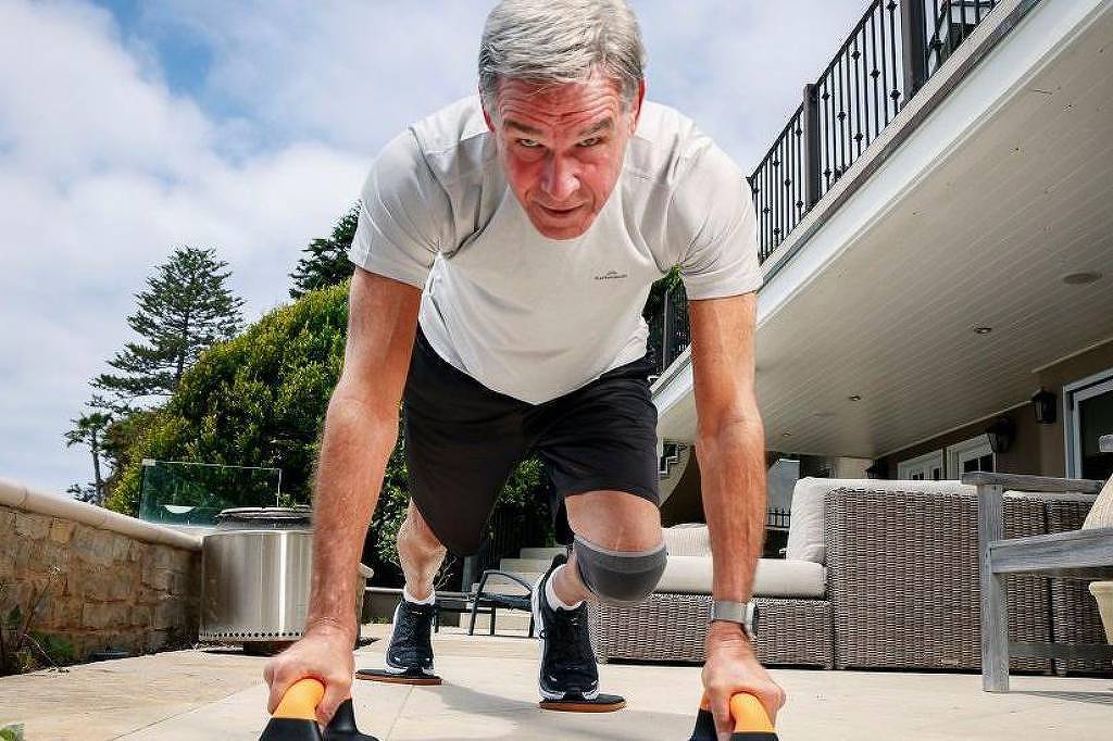 Um homem de cabelos grisalhos está realizando um exercício de empurrar no chão, usando equipamentos de treino. Ele está vestido com uma camiseta branca e shorts pretos. O ambiente é ao ar livre, com uma casa ao fundo e móveis de jardim visíveis. O céu está parcialmente nublado e há árvores ao fundo.