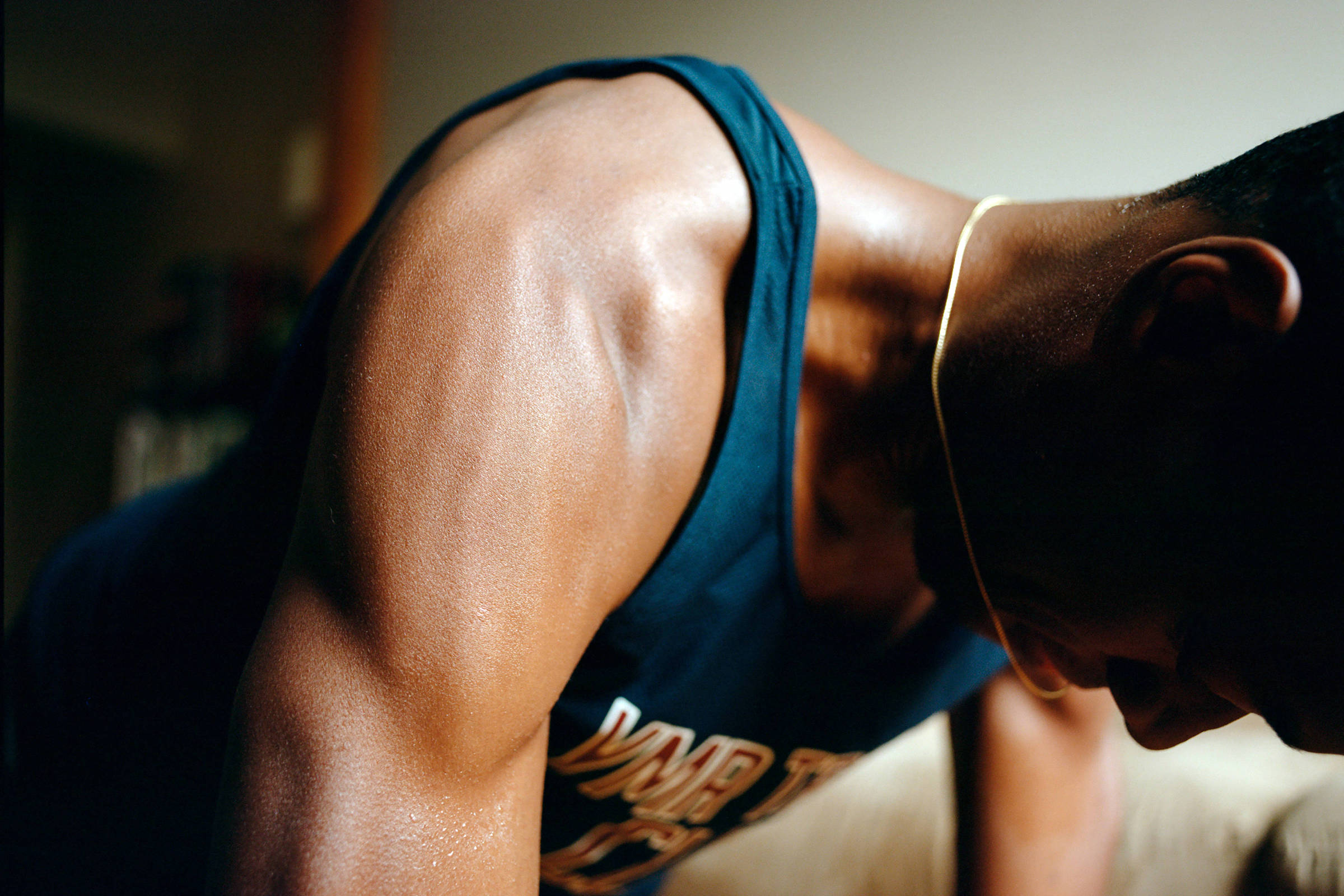 A imagem mostra um homem em posição de flexão, com o foco em seu braço e parte superior do corpo. Ele está usando uma camiseta sem mangas e parece estar suado, indicando esforço físico. O fundo é desfocado, mas sugere um ambiente interno, possivelmente uma sala de estar.