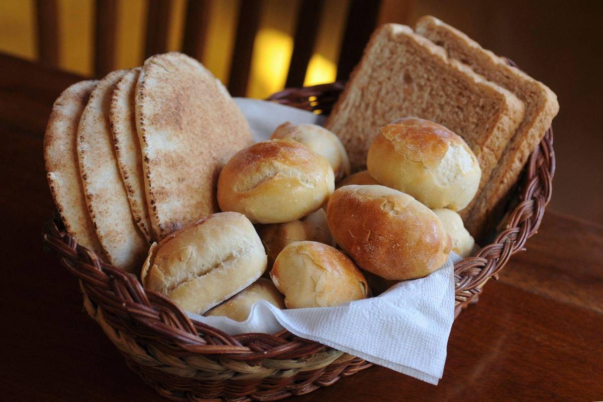 Uma cesta de pães contendo pães redondos e fatias de pão de forma. Os pães redondos estão dispostos em um pano branco dentro da cesta, enquanto as fatias de pão de forma estão empilhadas ao fundo. A cesta é feita de vime e está sobre uma superfície de madeira.