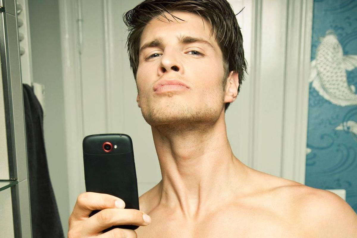 Um homem jovem está tirando uma selfie em um banheiro. Ele está sem camisa e segurando um celular com a mão direita. O fundo apresenta uma parede decorada com um padrão de peixes e há um espelho refletindo parte do ambiente. O homem tem cabelo castanho claro e está fazendo uma expressão confiante.