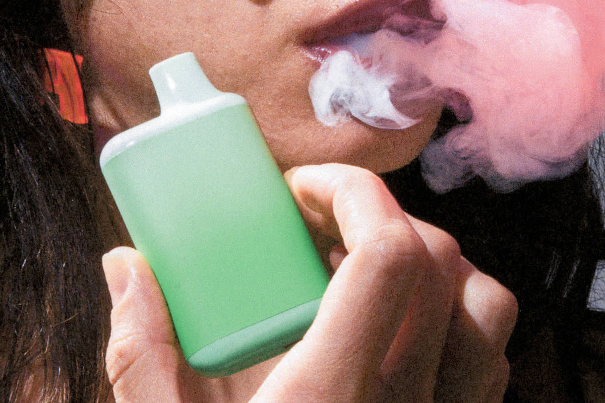 A imagem mostra uma pessoa segurando um dispositivo de vaporização verde, enquanto exala uma nuvem de vapor. O foco está na mão que segura o dispositivo e na fumaça que sai da boca da pessoa. O fundo é desfocado, destacando o ato de vaporização.