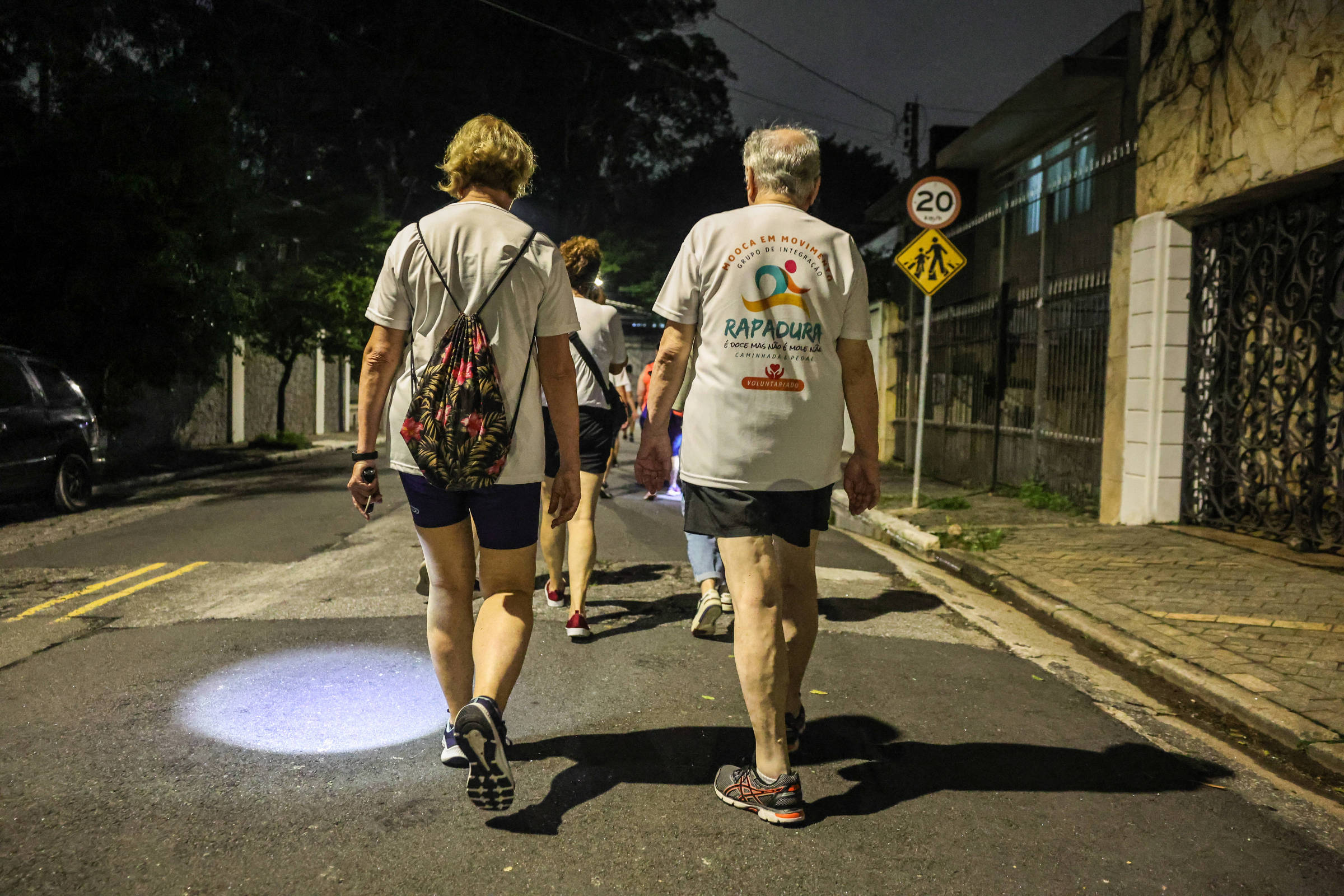 Um grupo de pessoas idosas caminha em uma rua à noite. Eles estão vestidos com camisetas claras e shorts, e um deles carrega uma mochila. A iluminação é baixa, e há uma luz no chão, possivelmente de uma lanterna. Ao fundo, há uma placa de trânsito indicando o limite de velocidade de 20 km/h e uma cerca ao lado da calçada.