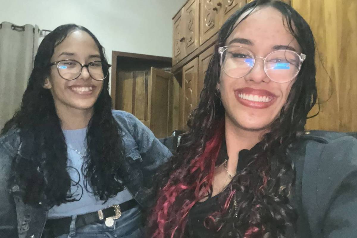 A imagem mostra duas jovens sorrindo para a câmera. Ambas têm cabelos longos e cacheados, uma delas com mechas vermelhas. Elas estão sentadas em um ambiente interno, com armários de madeira ao fundo. A jovem à esquerda usa óculos e uma jaqueta jeans, enquanto a outra está vestindo uma blusa preta. Ambas parecem felizes e estão em um momento descontraído.