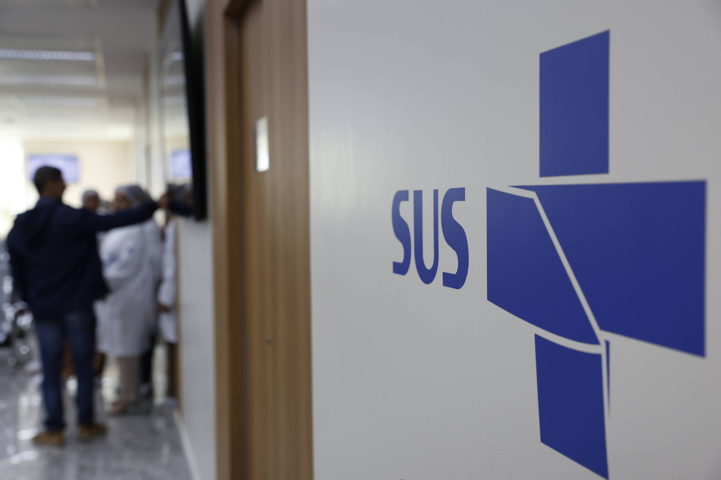 A imagem mostra uma parede com o logotipo do SUS (Sistema Único de Saúde) em destaque, na cor azul. Ao fundo, é possível ver pessoas em um ambiente hospitalar, algumas vestindo jalecos brancos, indicando que são profissionais de saúde. A cena sugere um ambiente de atendimento médico.