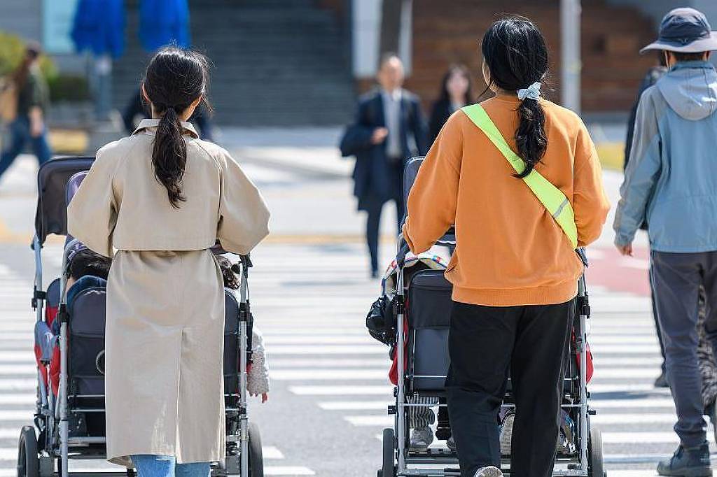 A imagem mostra duas mulheres caminhando em um cruzamento, cada uma empurrando um carrinho de bebê. A mulher à esquerda está vestida com um casaco claro, enquanto a mulher à direita usa um moletom laranja com uma faixa verde. Ao fundo, há pessoas atravessando a rua e um edifício visível.