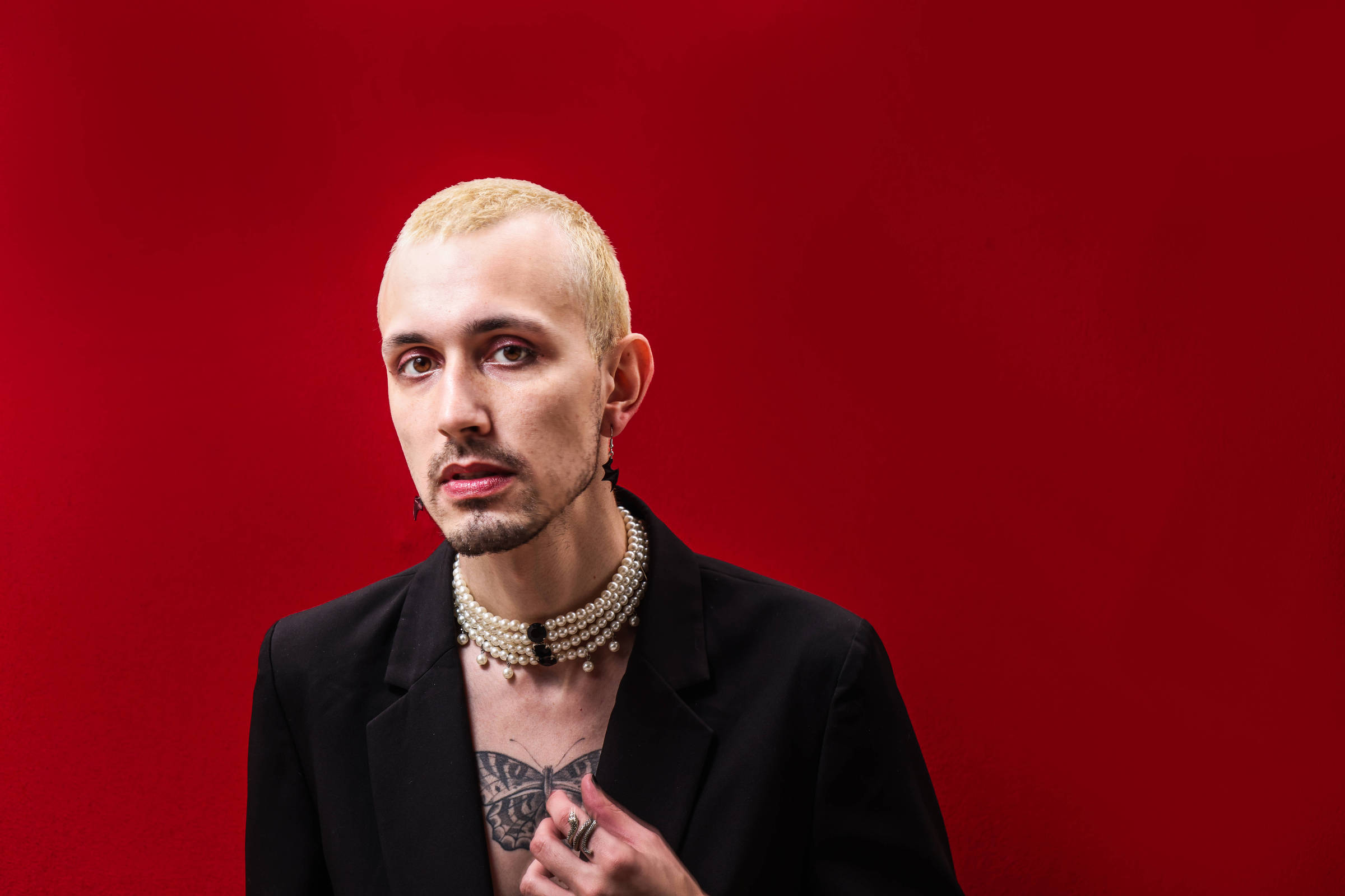 A imagem mostra uma pessoa com cabelo loiro platinado e pele clara, posando com as mãos levantadas. A pessoa usa um blazer preto e um colar de pérolas, além de várias anéis e esmalte nas unhas em cores diferentes, incluindo vermelho e preto. O fundo é uma parede branca texturizada.