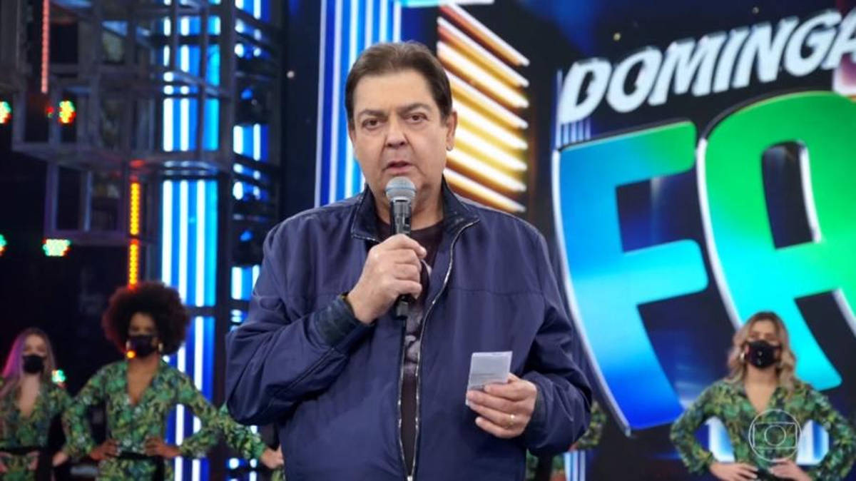 Um apresentador de televisão está em um palco, segurando um microfone e lendo um papel. Ele está vestido com uma jaqueta escura e tem cabelo castanho. Ao fundo, há uma tela grande com a palavra