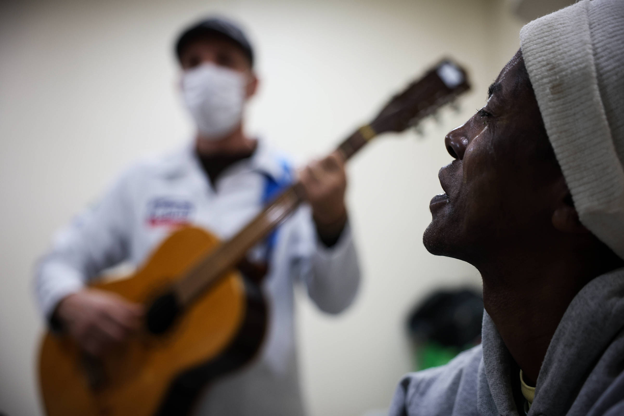 A imagem mostra um homem de pele escura, com um gorro branco, olhando atentamente para um músico ao fundo. O músico, que usa uma máscara facial e um chapéu, toca um violão. O ambiente parece ser uma sala interna, com uma lixeira visível ao fundo. A iluminação é suave, destacando a expressão do homem que escuta a música.
