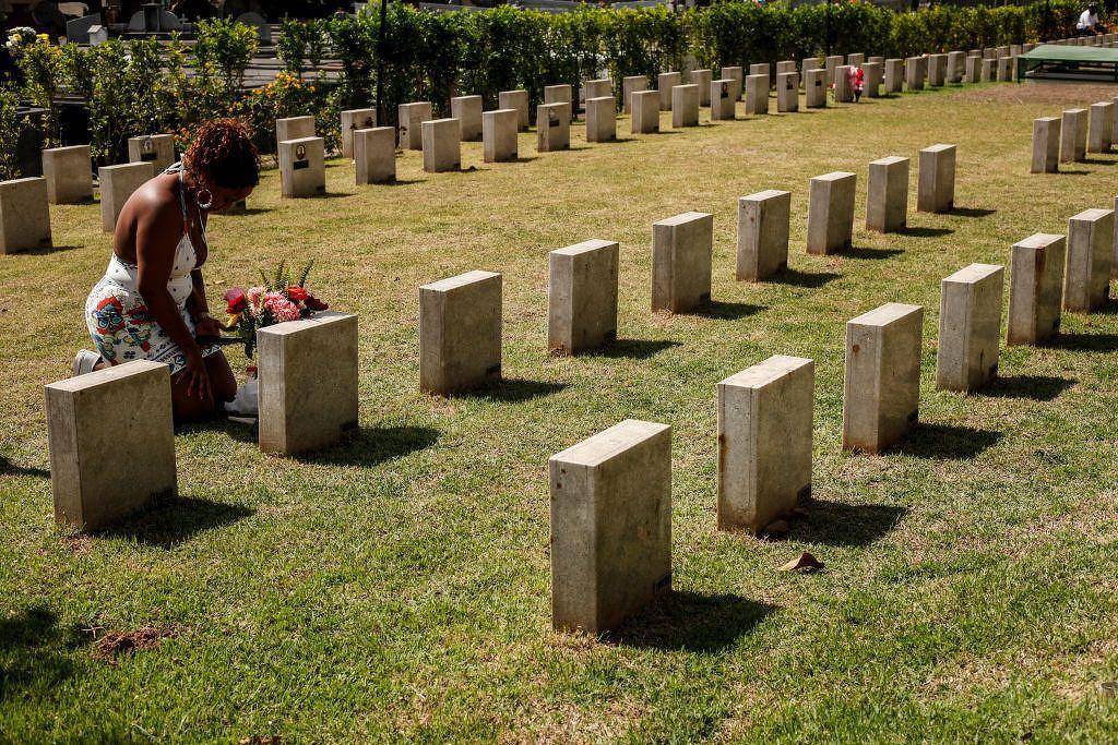 Uma mulher está ajoelhada em um cemitério, colocando flores em um dos túmulos. Ao fundo, há várias lápides retangulares dispostas em linhas, com grama verde ao redor. A mulher usa um vestido claro e está concentrada em sua homenagem. O ambiente é tranquilo e bem cuidado, com arbustos ao fundo.