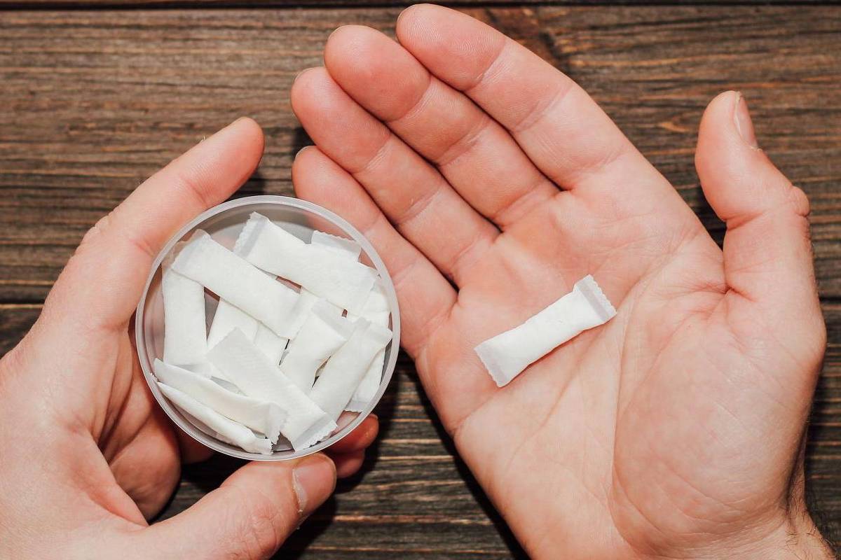 Uma mão segura um recipiente transparente com várias pequenas porções brancas, enquanto a outra mão segura uma única porção. O fundo é uma superfície de madeira escura.