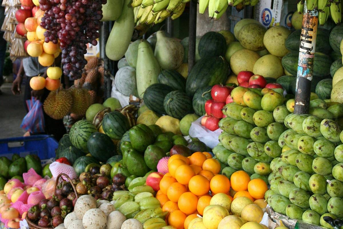 A imagem mostra uma banca de frutas em um mercado. Há uma variedade de frutas expostas, incluindo bananas amarelas penduradas, uvas roxas, melões, maçãs, laranjas e outras frutas de diferentes cores e tamanhos. A disposição das frutas é organizada, criando um visual vibrante e colorido.