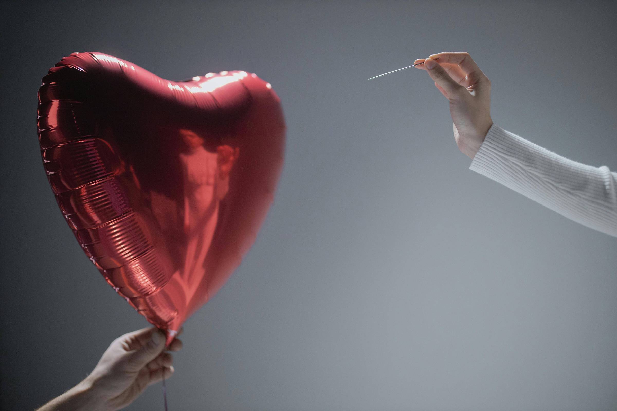 A imagem mostra um balão em forma de coração, vermelho e metálico, sendo segurado por uma mão à esquerda. À direita, outra mão está prestes a furar o balão com uma agulha. O fundo é neutro e desfocado, destacando o balão e as mãos.