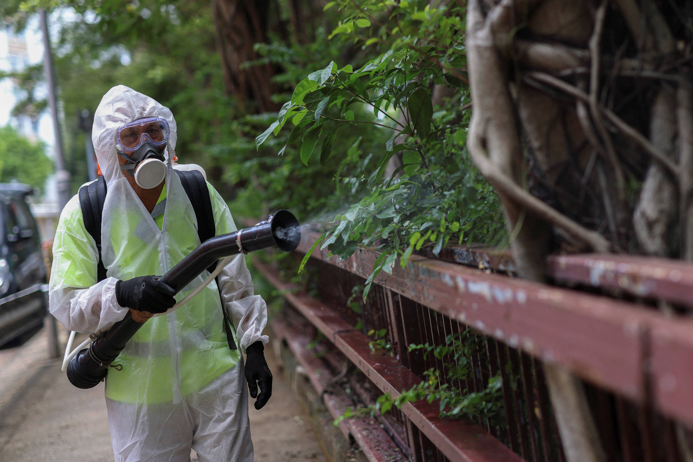Um trabalhador vestido com um traje de proteção, incluindo uma máscara e óculos, está aplicando inseticida em uma área verde ao lado de uma cerca. Ele utiliza um equipamento de pulverização para tratar as plantas e arbustos ao redor. O ambiente é urbano, com árvores e vegetação ao fundo.
